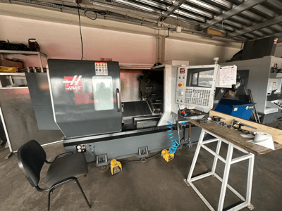Tornio CNC Haas (modello ST-10) in un'officina, vista laterale che mostra i comandi, gli utensili su un banco di lavoro e una sedia nelle vicinanze.