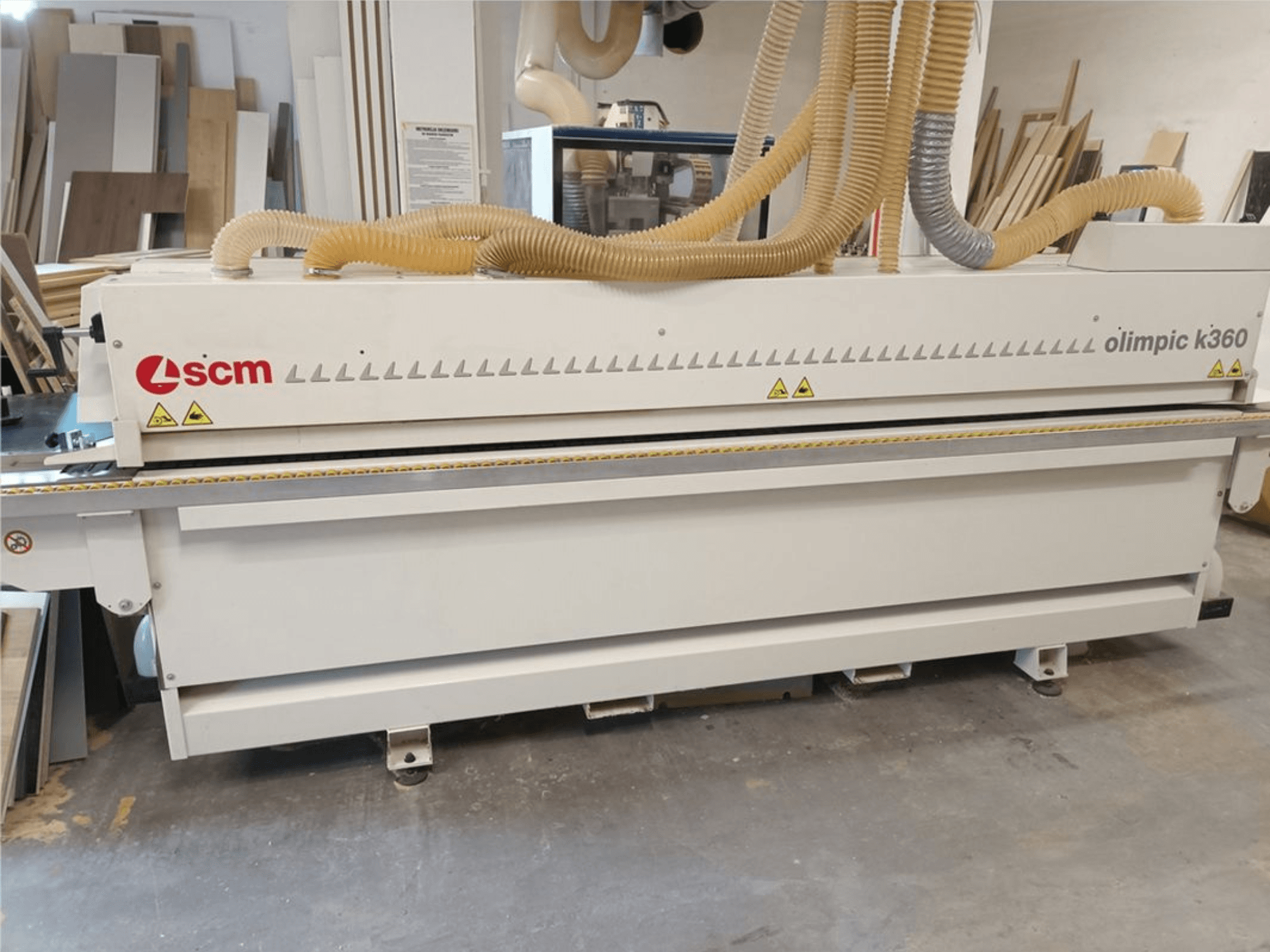 Bordatrice SCM Olimpic K360 T-ER1 in un'officina, vista frontale che mostra i tubi di aspirazione della polvere e le etichette di avvertenza.