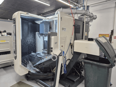 Fresatrice DMG aperta con spruzzi di refrigerante, che mostra trucioli di metallo e comandi degli utensili, vista dal davanti.