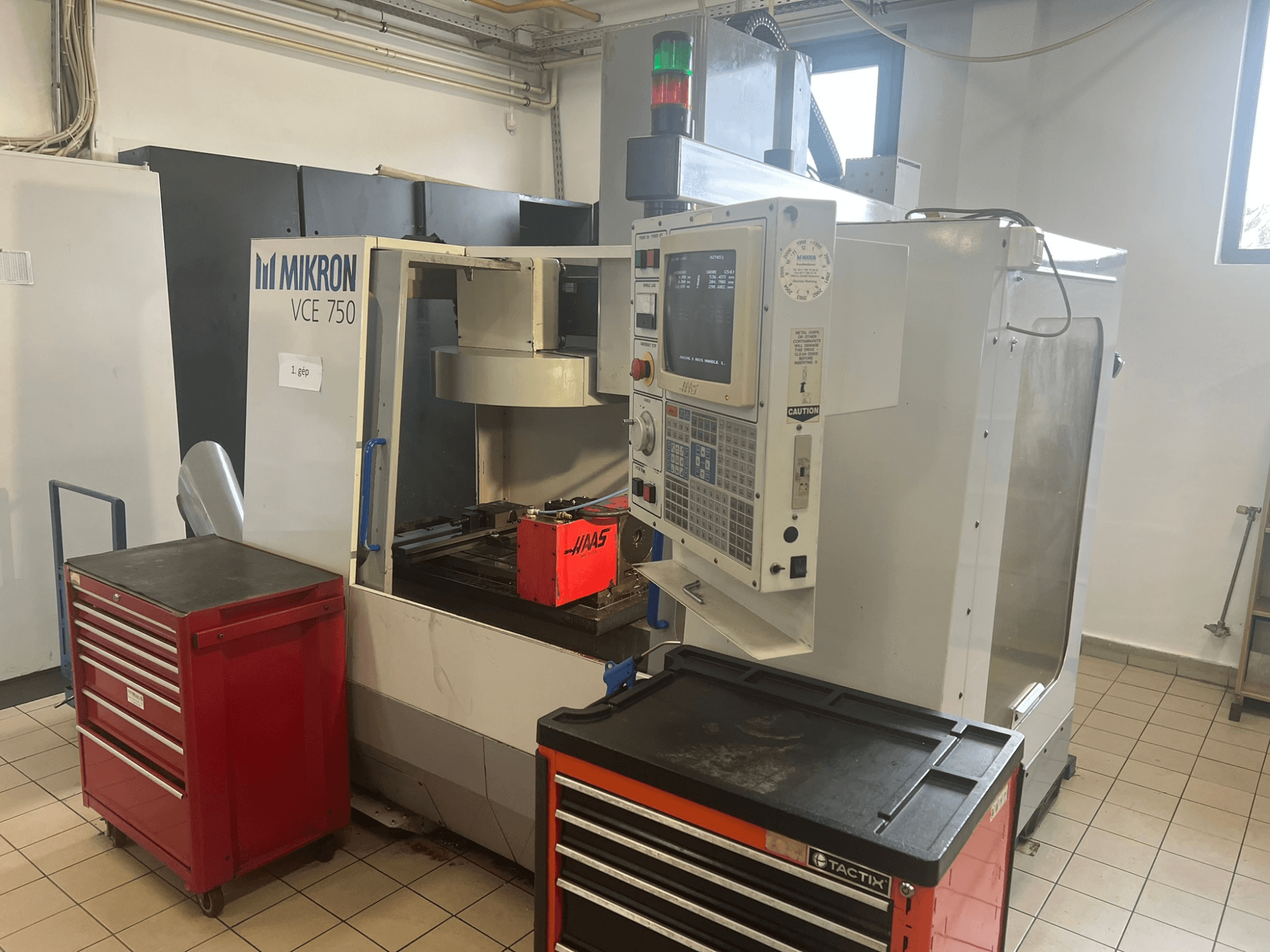 Testo Alt: "Centro di lavoro verticale MIKRON VCE 750, vista frontale con pannello di controllo e cassette degli attrezzi rosse, in un'ampia officina".