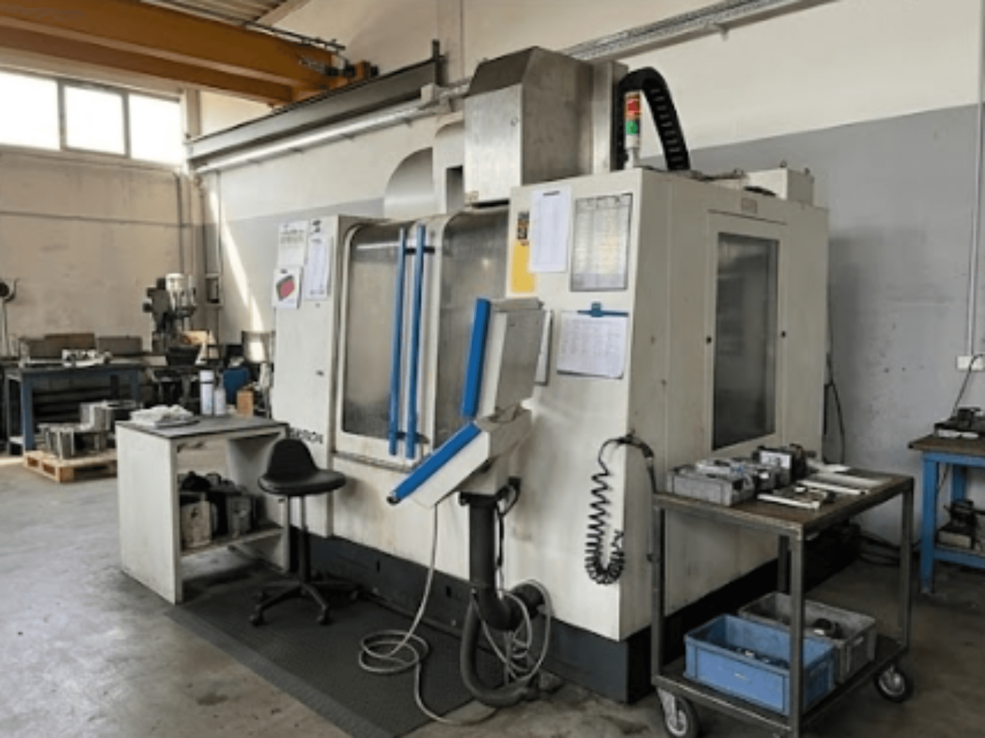 Centro di lavorazione verticale MIKRON VCE 1000 Pro in un'officina, vista frontale con dettagli blu e piano di lavoro adiacente.