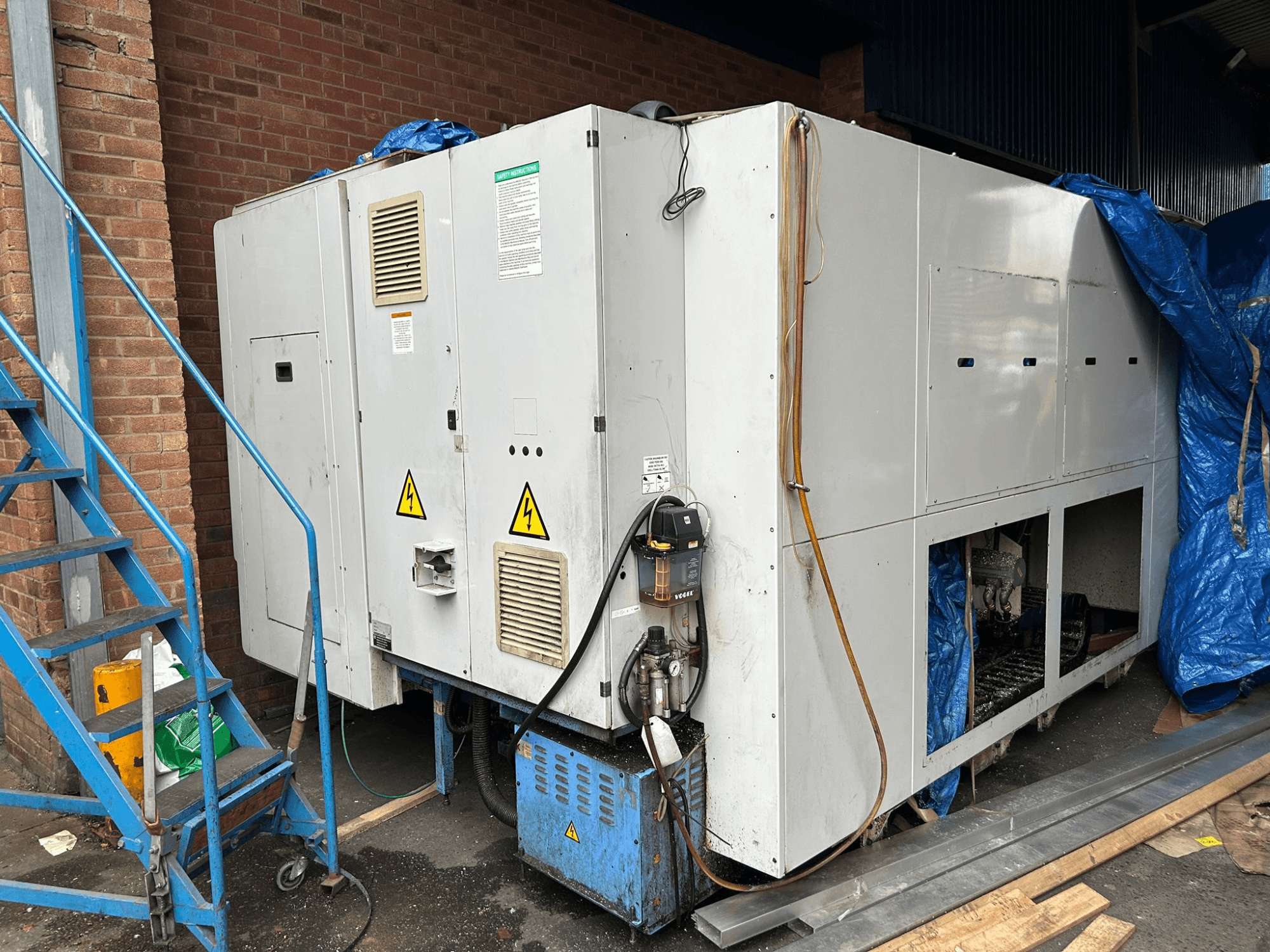 Vista laterale di un grande refrigeratore industriale, marca e modello non chiari, che mostra i collegamenti elettrici, i cartelli di avvertimento e il telo di copertura blu.