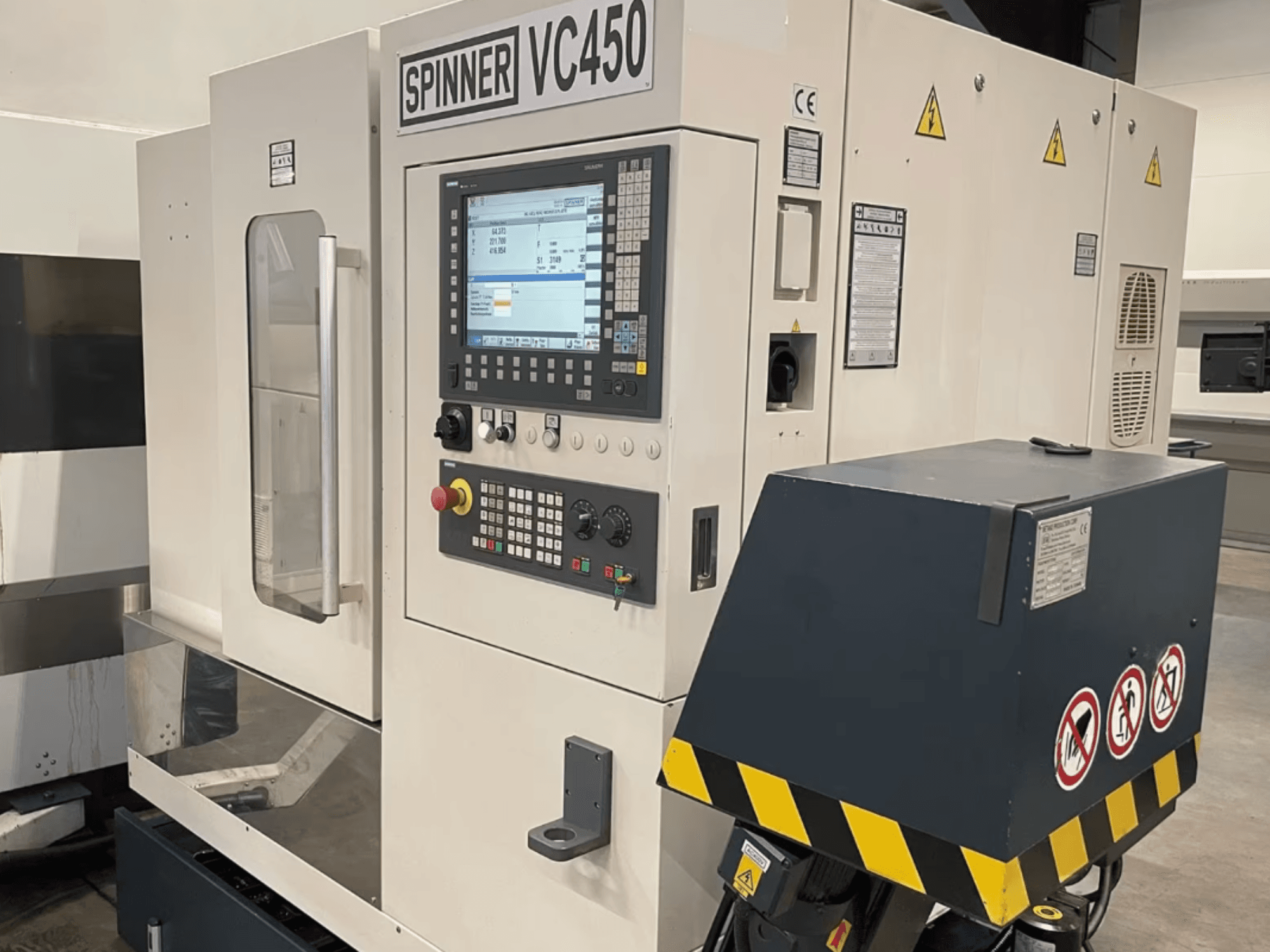 Vista frontale del centro di lavorazione verticale SPINNER VC450, con pannello di controllo ed etichette di sicurezza, che ne illustra i dettagli operativi.