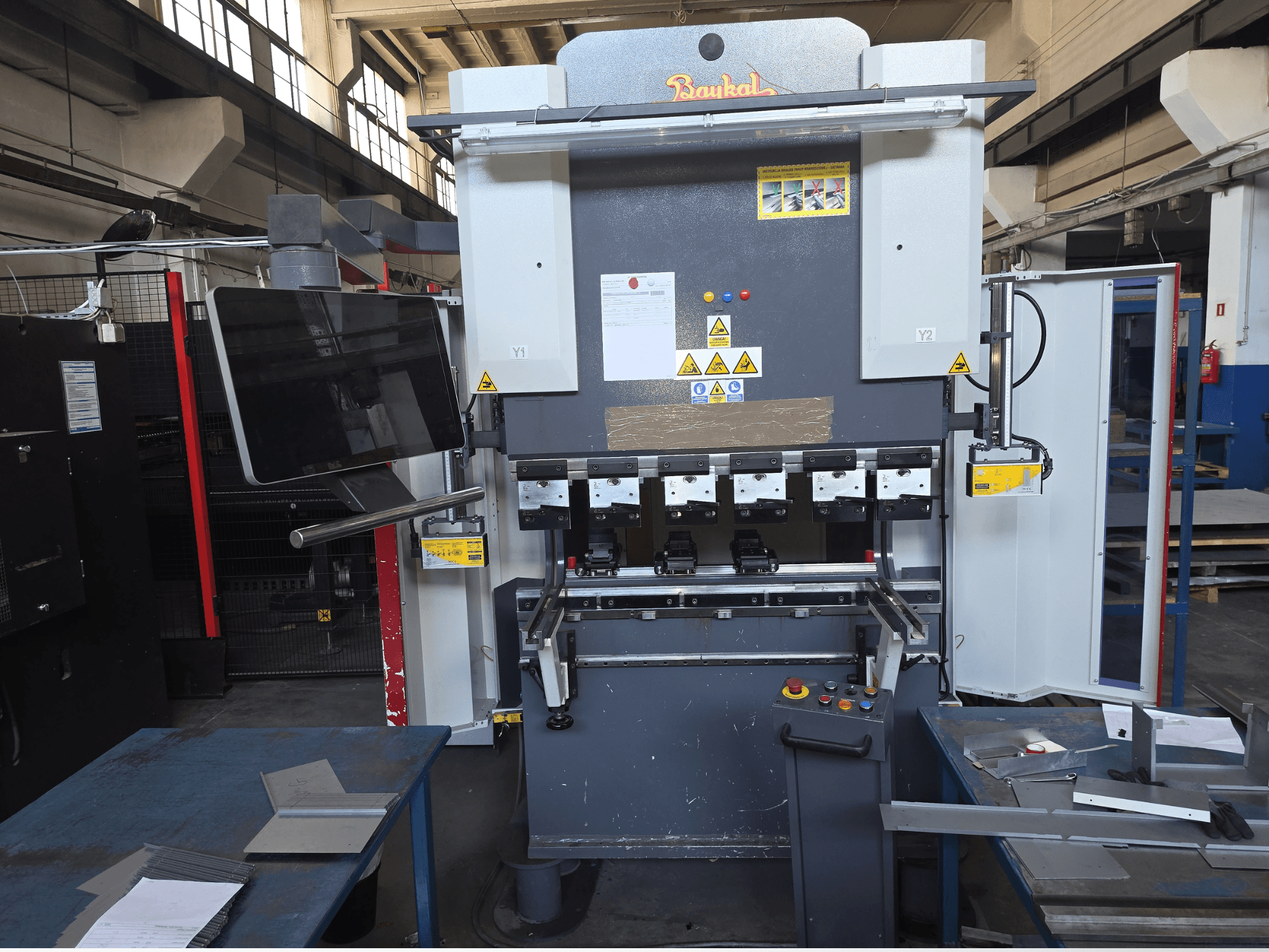 Piegatrice Baykal APHS 125040, vista frontale, che mostra il pannello di controllo e i dispositivi di sicurezza con i materiali su un tavolo da lavoro blu.