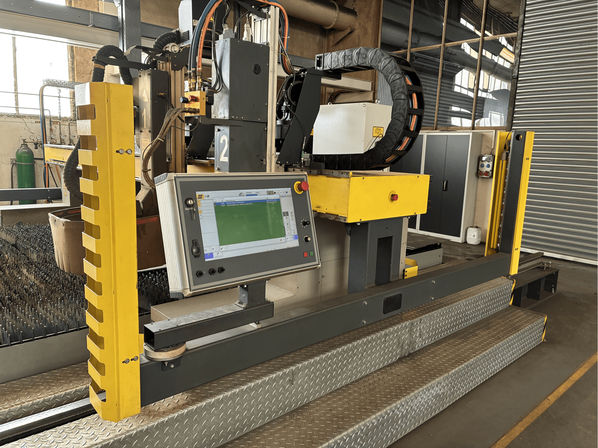 Macchina da taglio industriale con pannello di controllo touchscreen, protezione di sicurezza gialla, vista frontale, base a griglia metallica visibile.