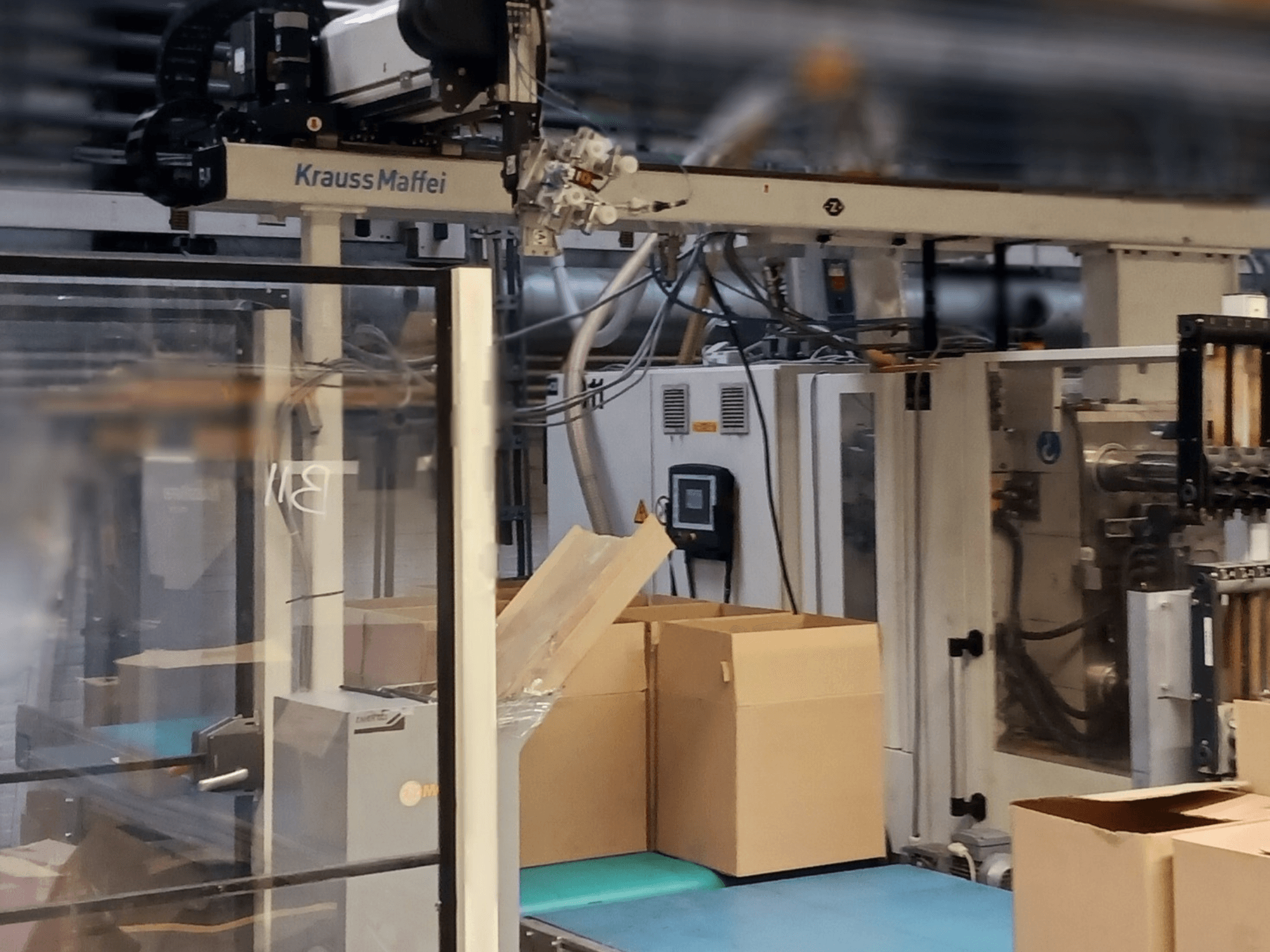 Sistema di automazione Krauss Maffei in una fabbrica, vista frontale che mostra scatole su un trasportatore e componenti del macchinario in funzione.