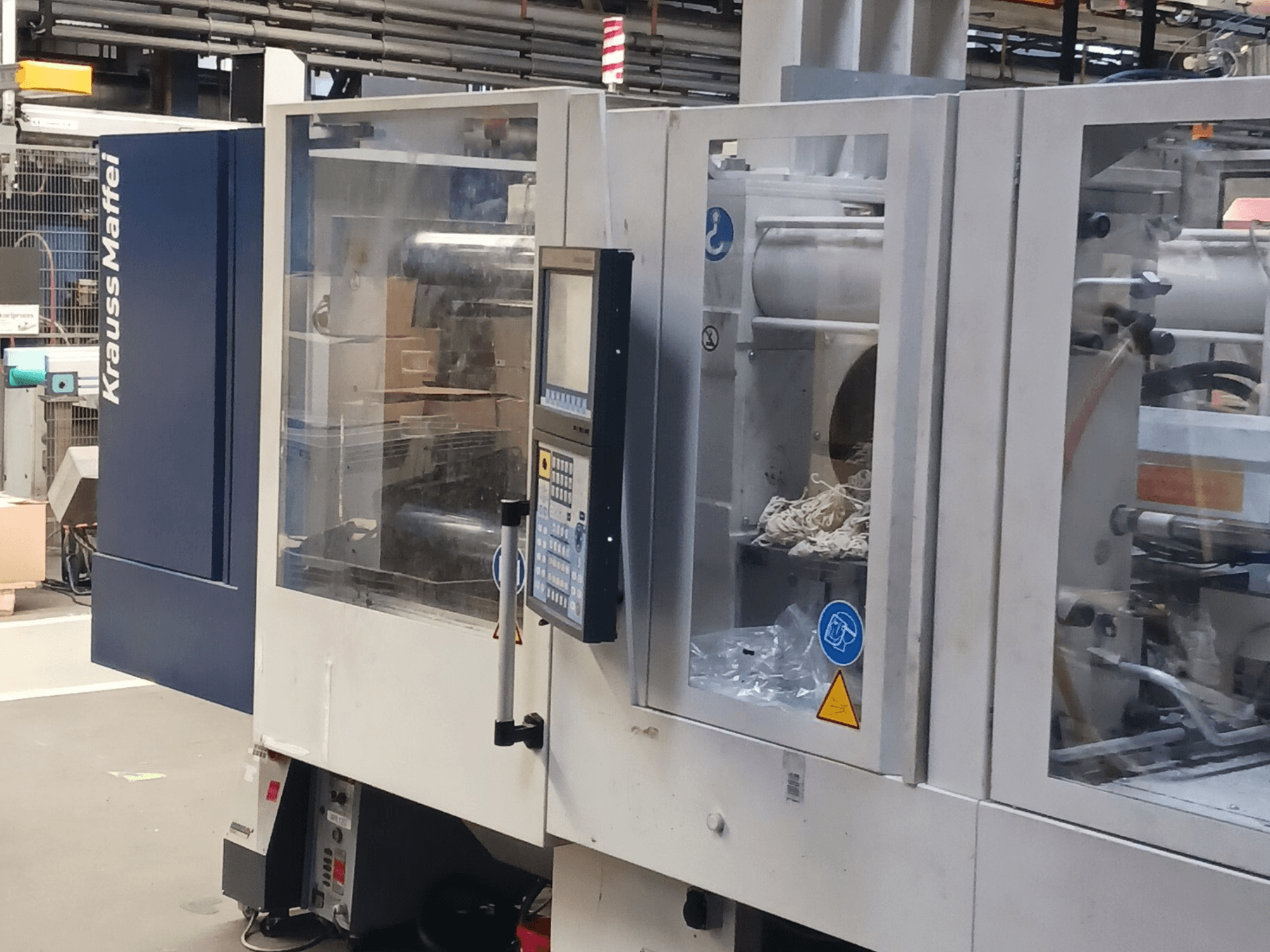 Macchina ad iniezione KraussMaffei, vista laterale, con pannello di controllo, macchinari e materiali di imballaggio sullo sfondo.