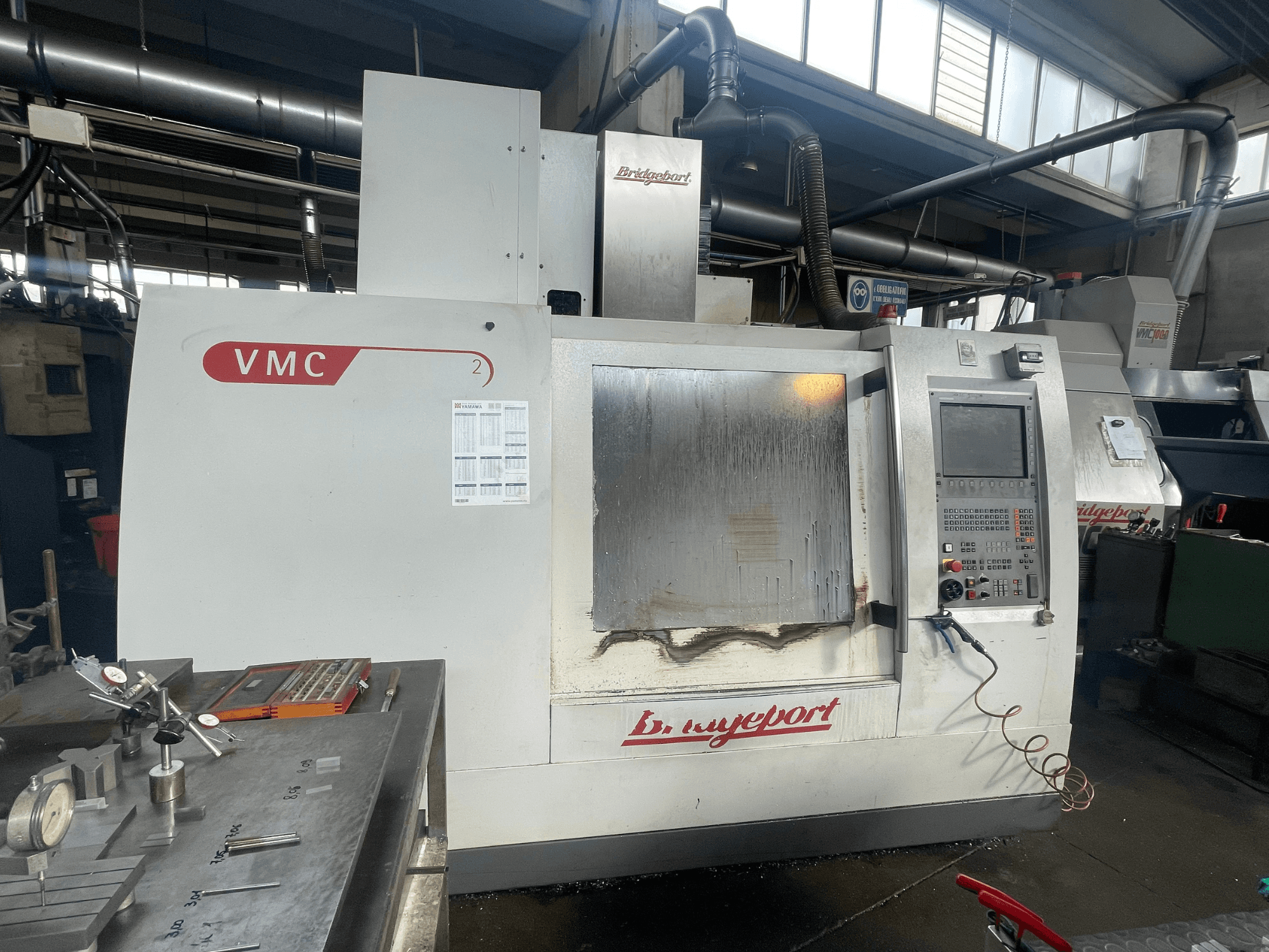 Centro di lavorazione verticale Bridgeport VCM 800-30 in un'officina, dotato di pannello di controllo e tavola portautensili. Vista frontale con dettagli visibili.