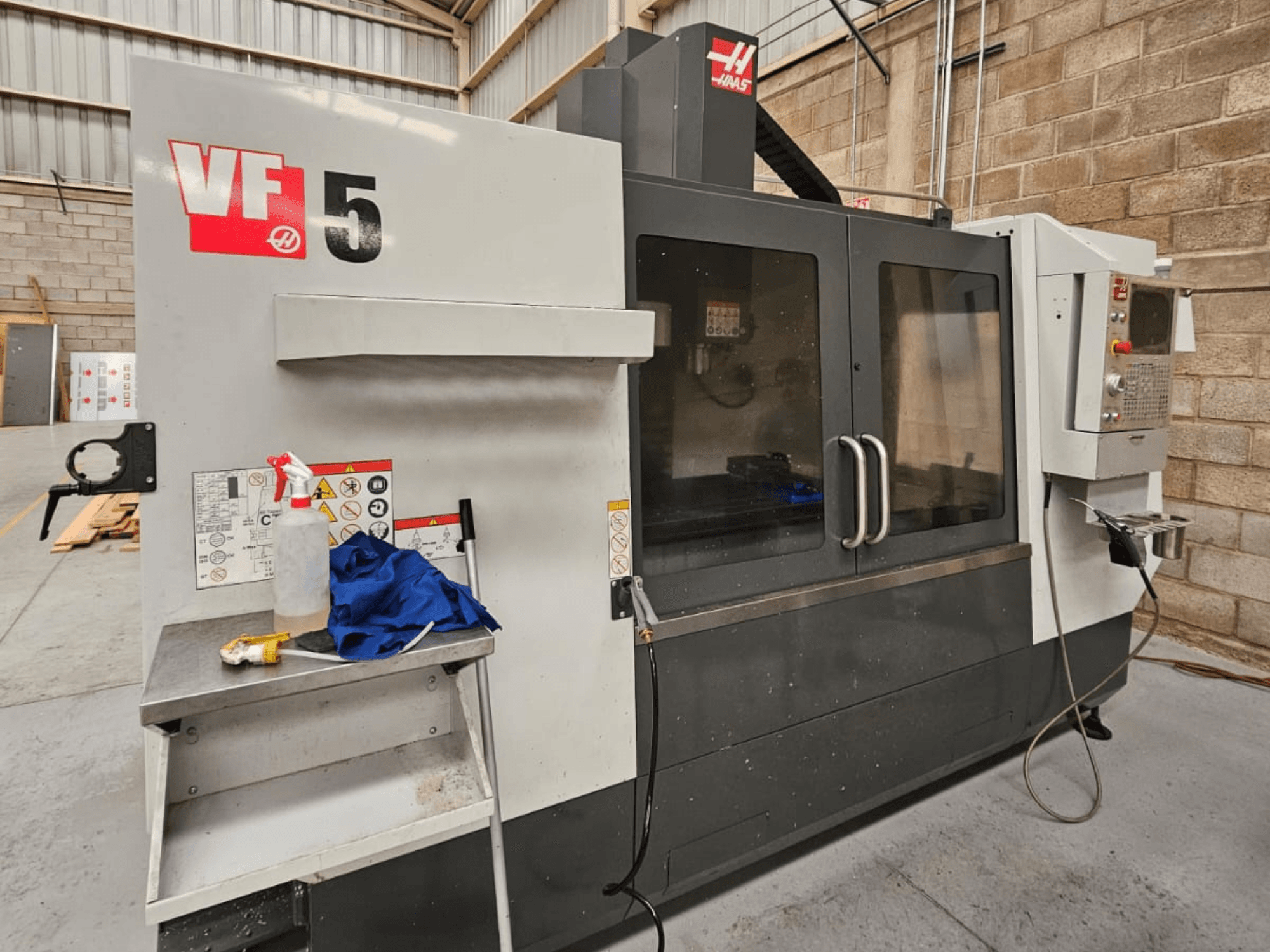 Fresatrice CNC Haas VF-5 in vista frontale, con pannello di controllo, bottiglia di refrigerante, panno blu e istruzioni di sicurezza.