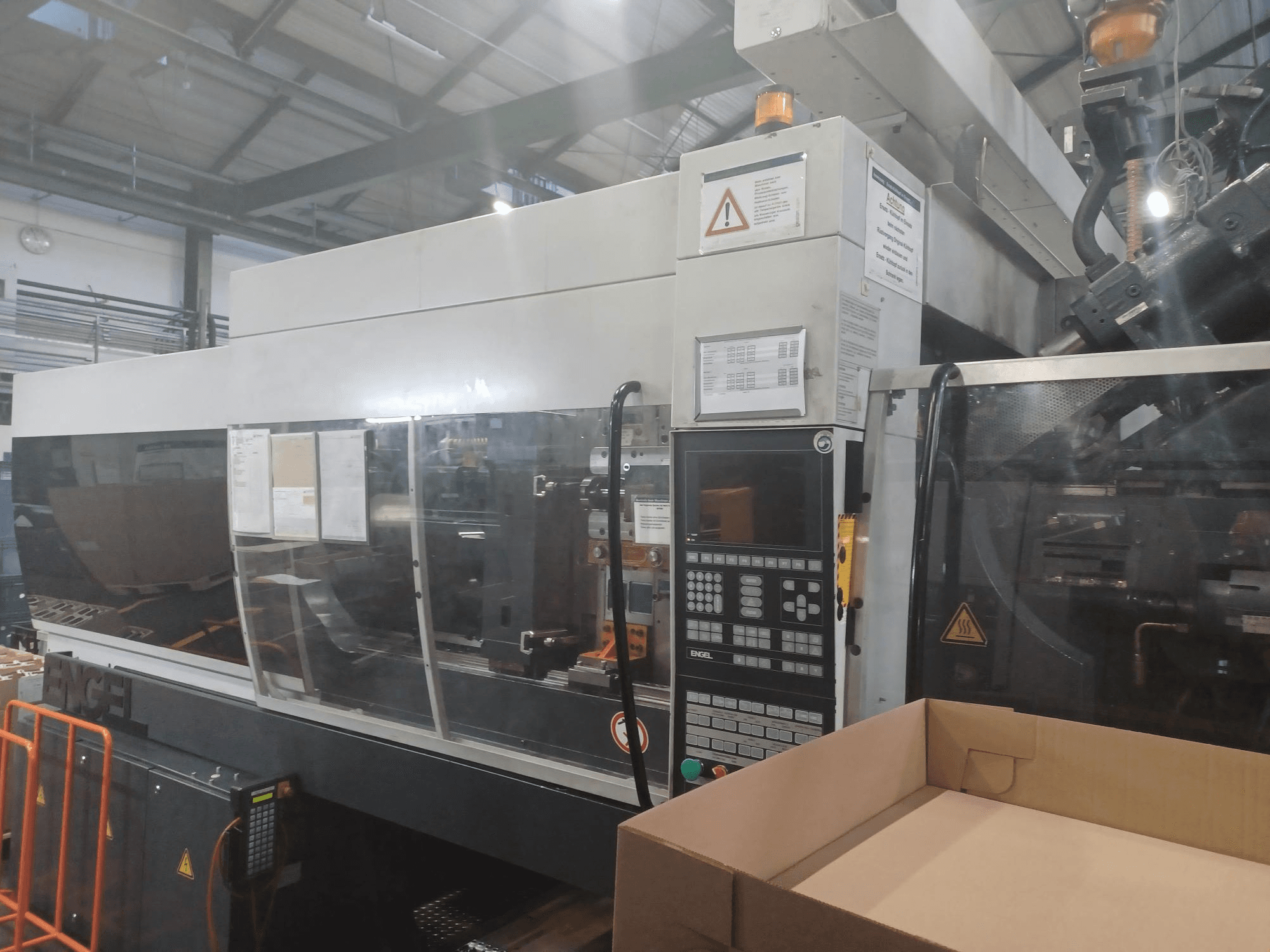 Macchina ad iniezione Engel, vista frontale, con pannello di controllo, etichette di sicurezza e una scatola vuota posizionata nelle vicinanze.