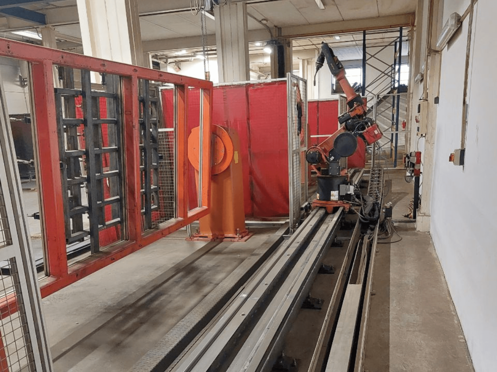 Braccio robotico industriale con colori arancioni e neri posizionato su binari metallici, circondato da barriere protettive rosse, vista frontale.