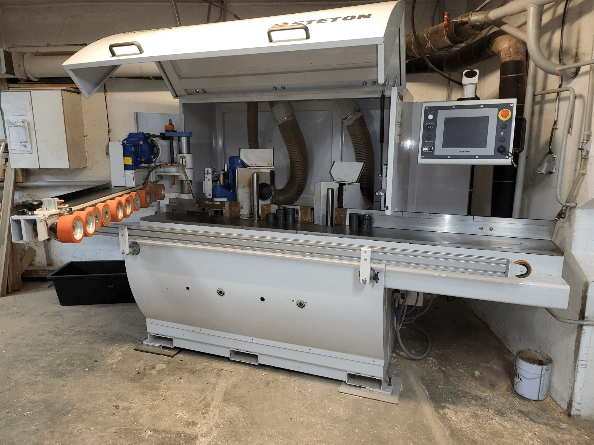 Vista frontale di una macchina per la lavorazione del legno Stetton con pannello di controllo, tubi di alimentazione e vari pezzi di legno sul tavolo.