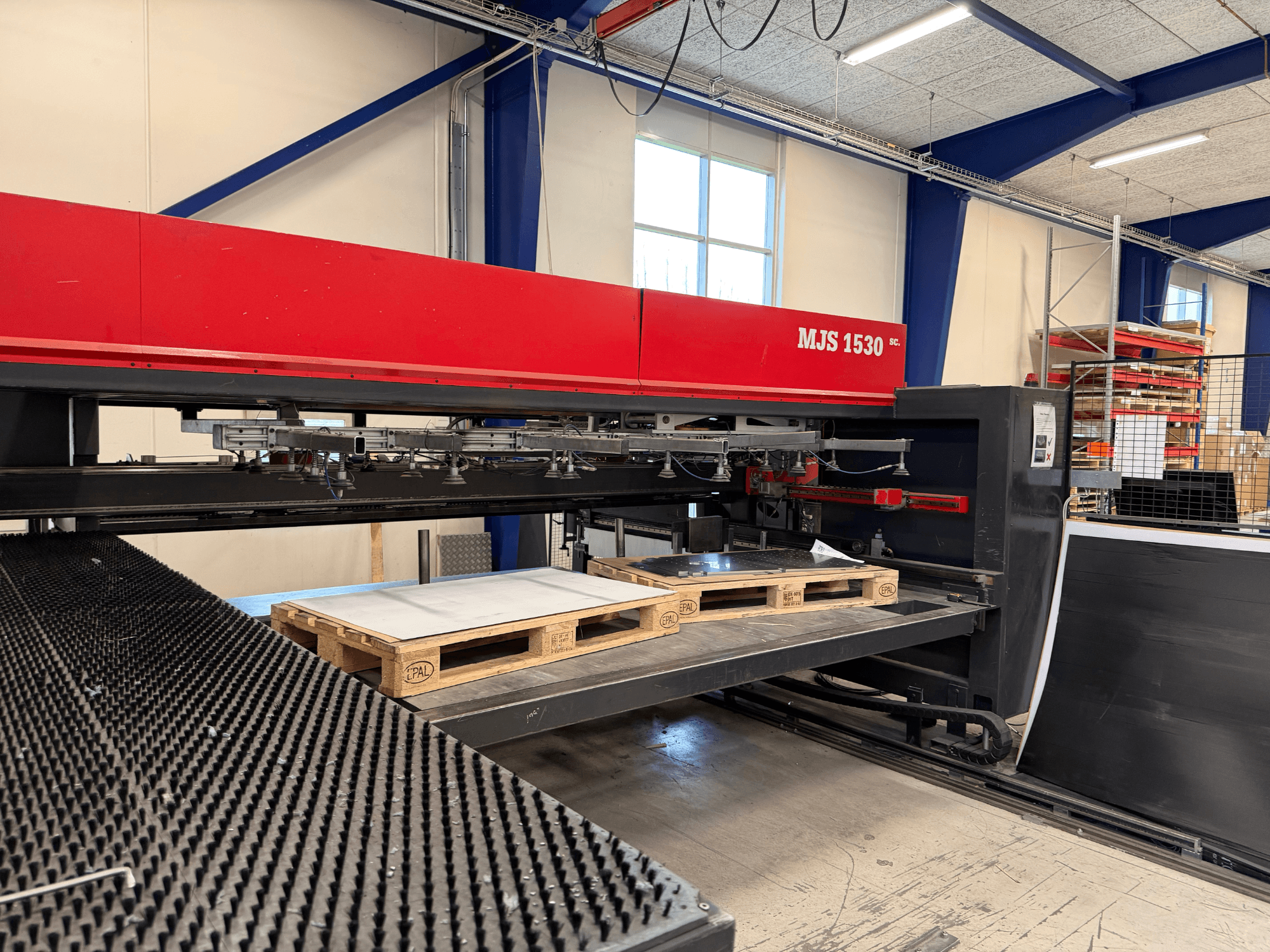 Macchina CNC MJS 1530 in un'officina, vista frontale, con un pallet di legno che sostiene una lamiera su una piattaforma rialzata.