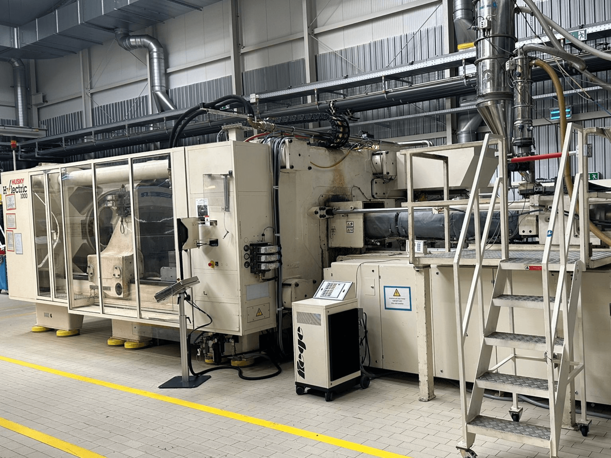 Vista frontale della pressa a iniezione elettrica Husky Hylelectric H1000 RS95/80 in un ambiente industriale, con pannello di controllo e gradini.