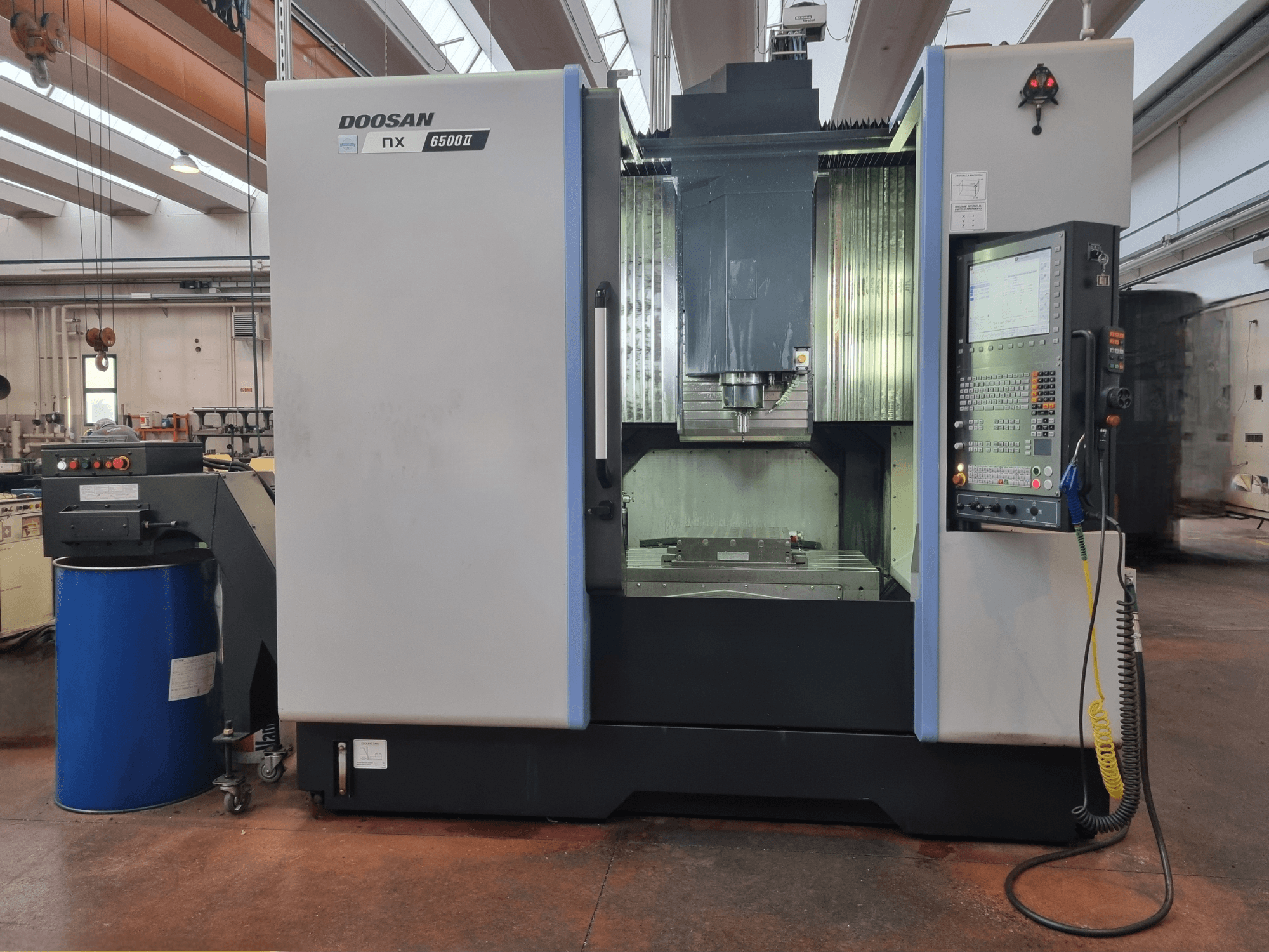 Macchina CNC Doosan NX 6500 II, vista frontale, con pannello di controllo e area di lavorazione, posizionata accanto a un barile blu.