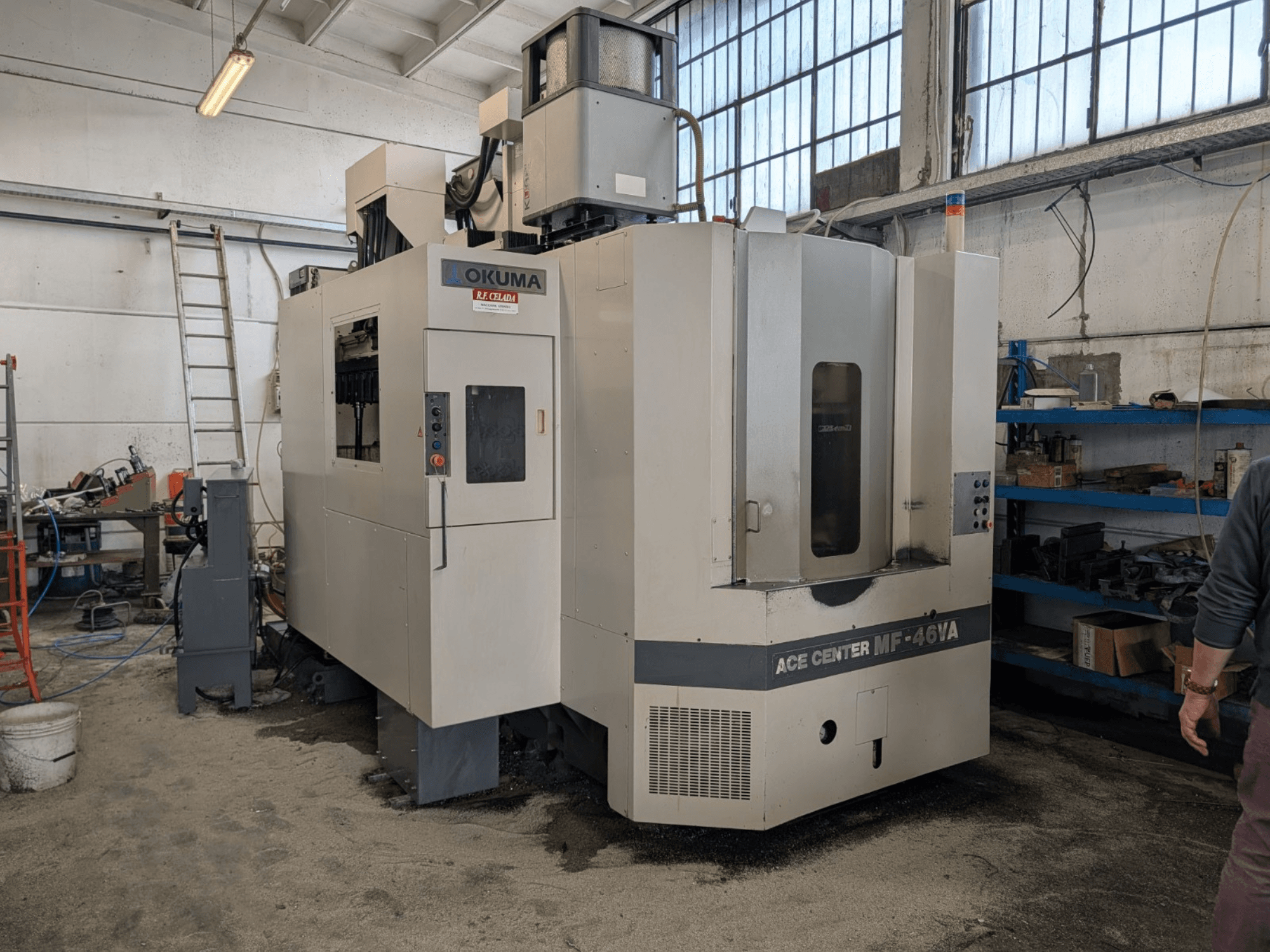 Centro di lavoro verticale Okuma ACE CENTER MF-46VA in un'officina, vista frontale con pannello di controllo e attrezzature circostanti.