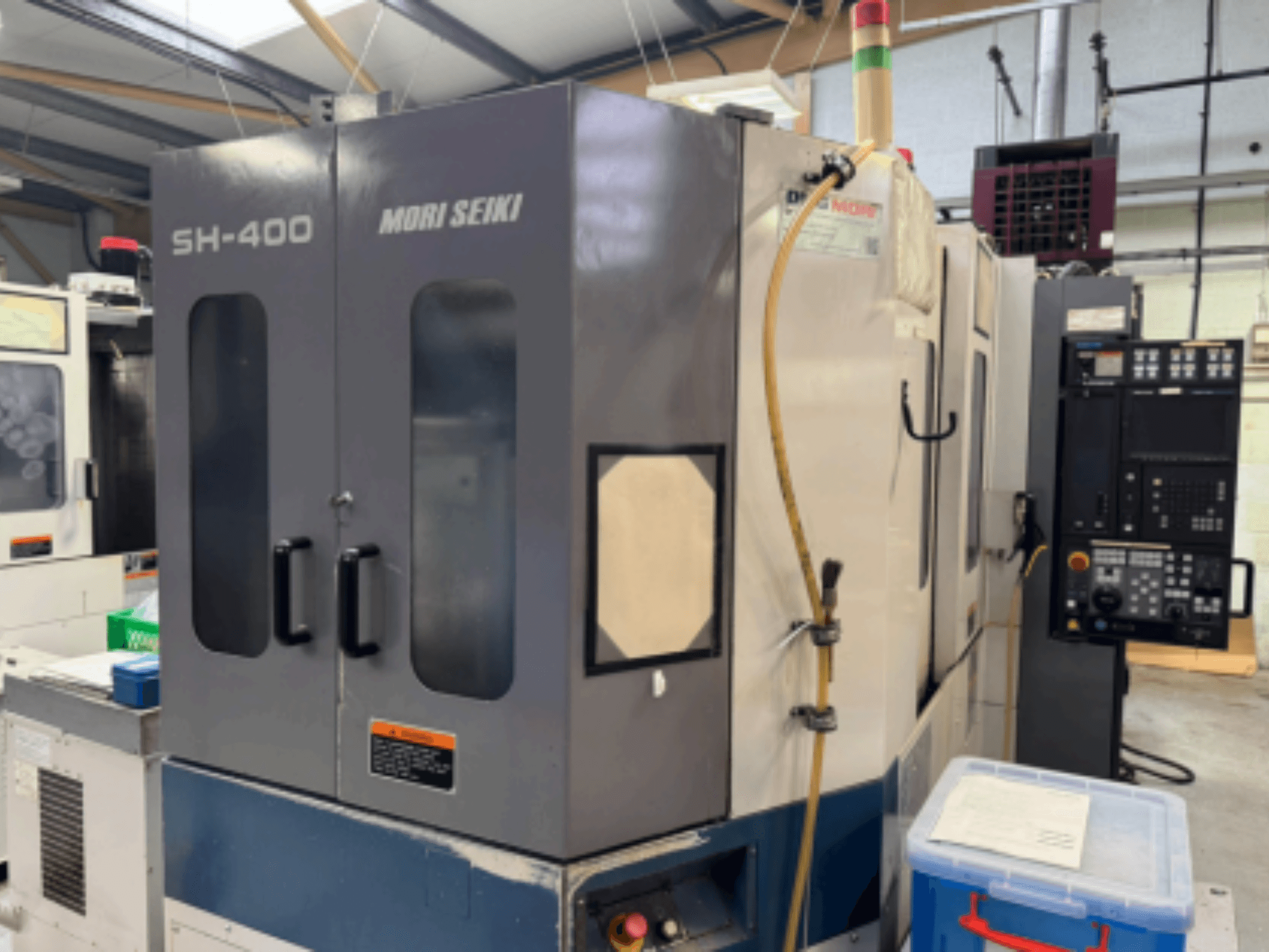 Macchina CNC Mori Seiki SH-400, vista frontale, con doppio sportello, pannello di controllo e tubo di collegamento. Contenitore di stoccaggio blu nelle vicinanze.