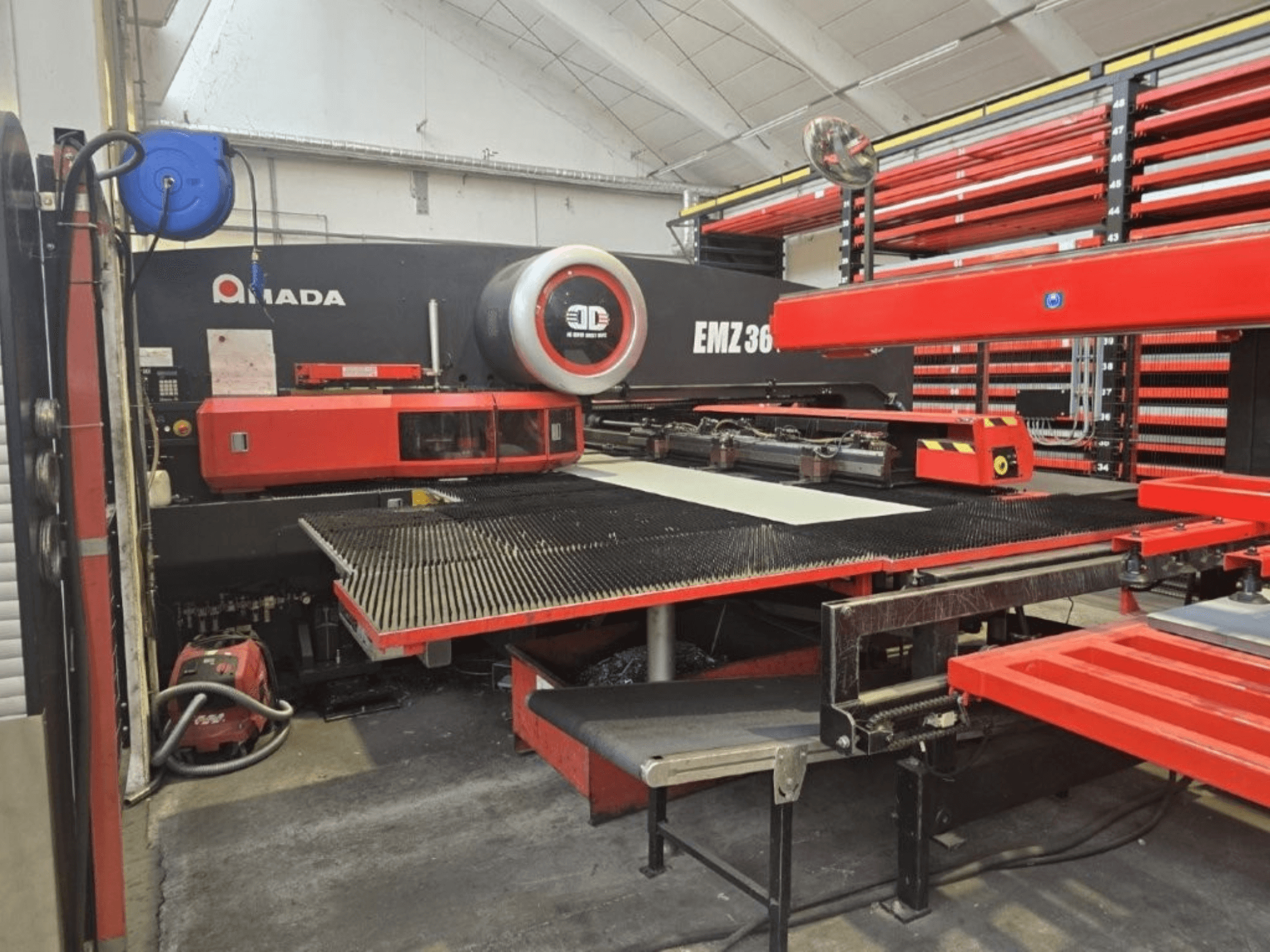 Macchina da taglio laser Amada EMZ 36 in un'officina, vista frontale, con accenti rossi e un grande tavolo da taglio.