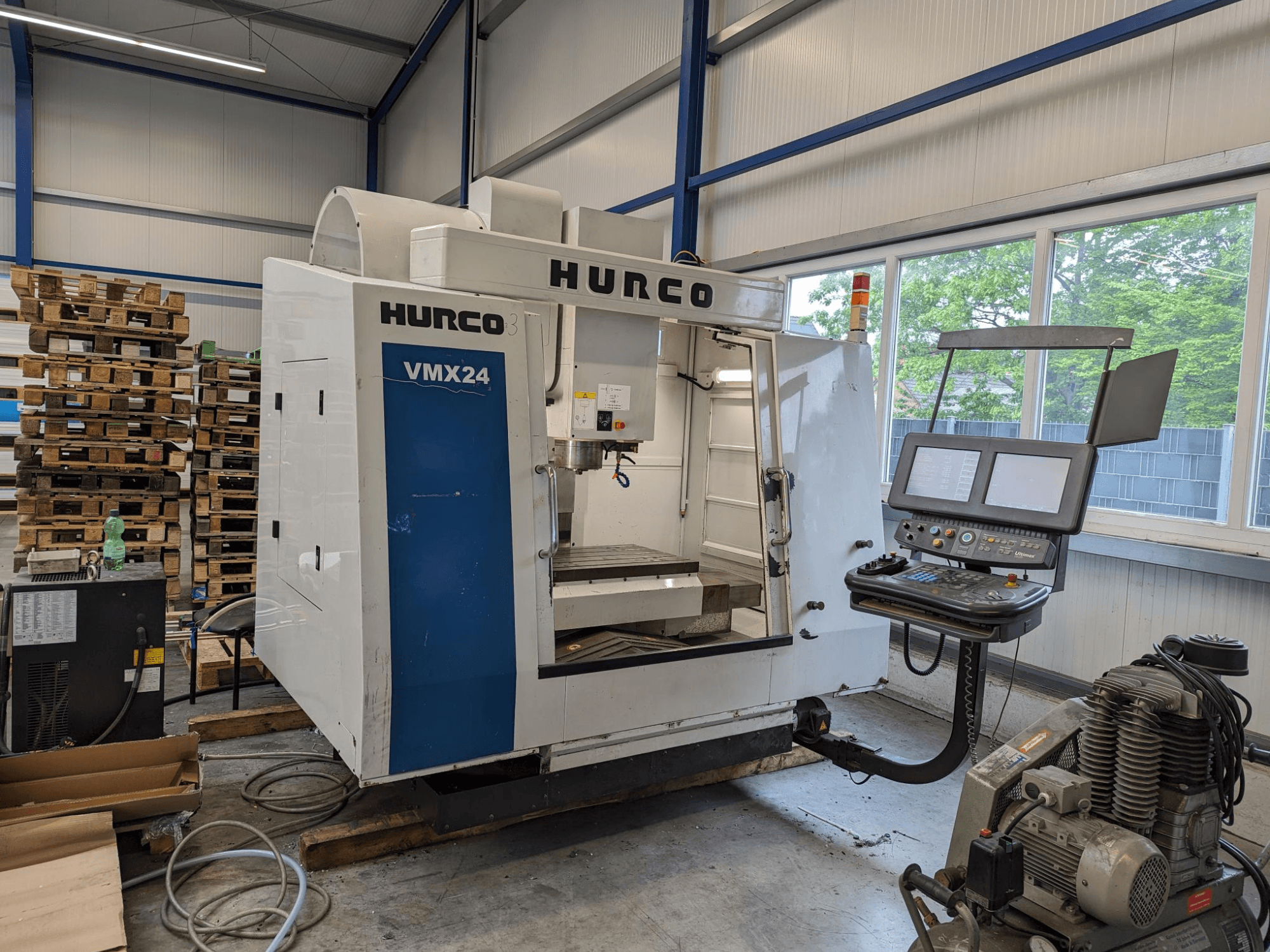 Fresatrice Hurco VMX24, vista frontale, con pannello di controllo e area utensili; sullo sfondo pallet impilati; ambiente di lavoro industriale.