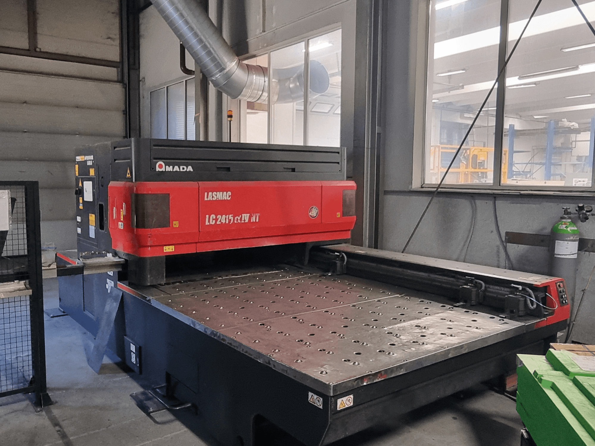 Macchina da taglio laser Amada LASMAC LC 2415 αⅡ NT in rosso e nero, vista frontale, con banco di taglio motorizzato e comandi di precisione.