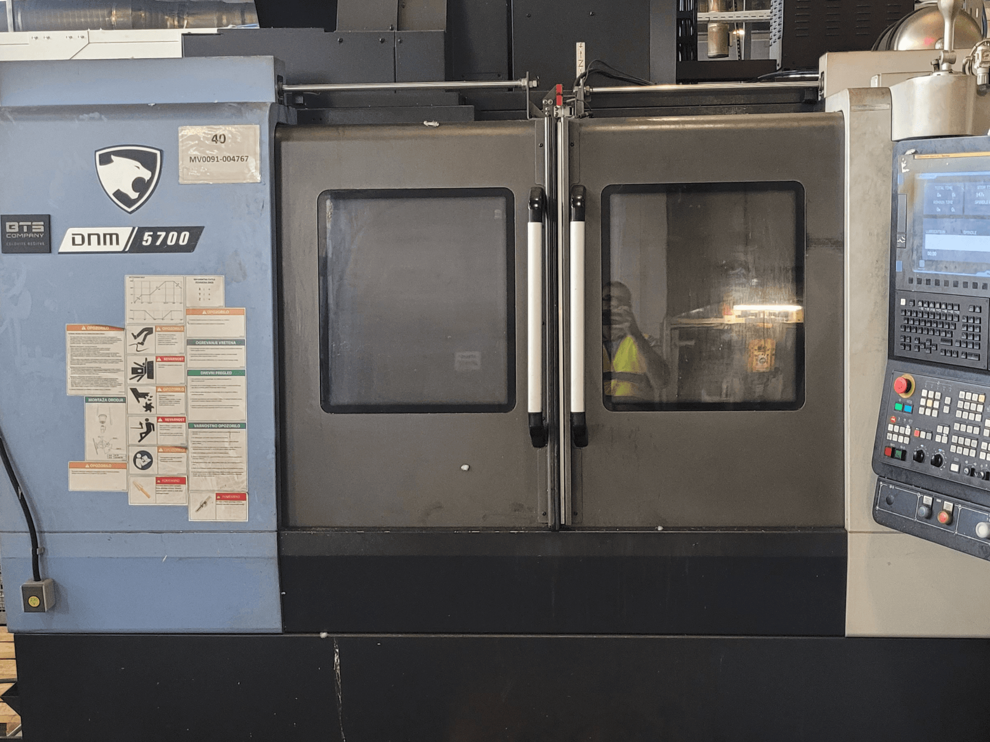 Vista frontale del centro di lavorazione verticale DOOSAN DNM 5700 con pannello di controllo, etichette di sicurezza e sportelli leggermente socchiusi.