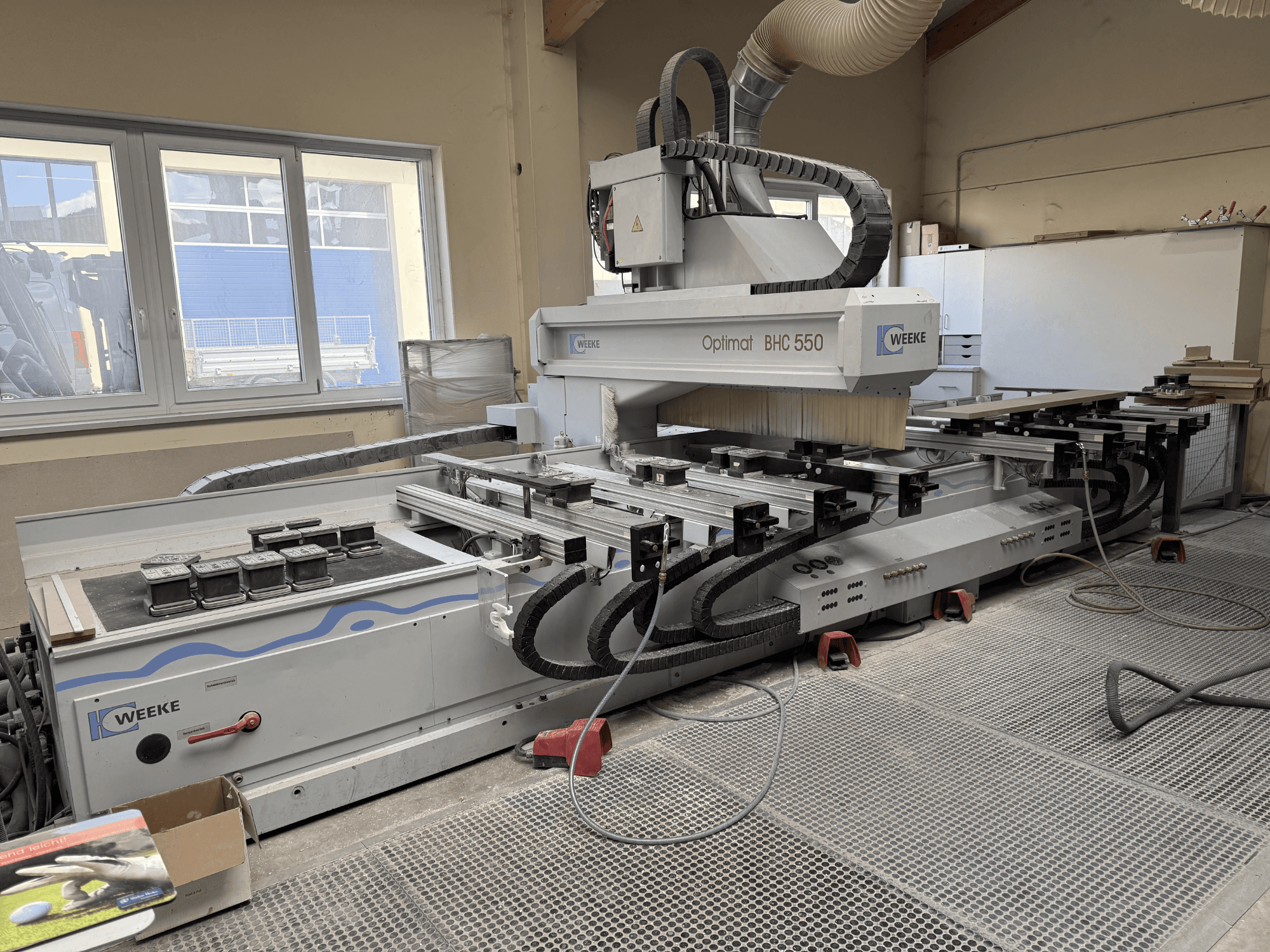 Centro di lavorazione del legno a CNC Weeke Optimat BHC 550, vista frontale che mostra la testa dell'utensile e il piano di lavoro, con sistema di aspirazione delle polveri visibile.