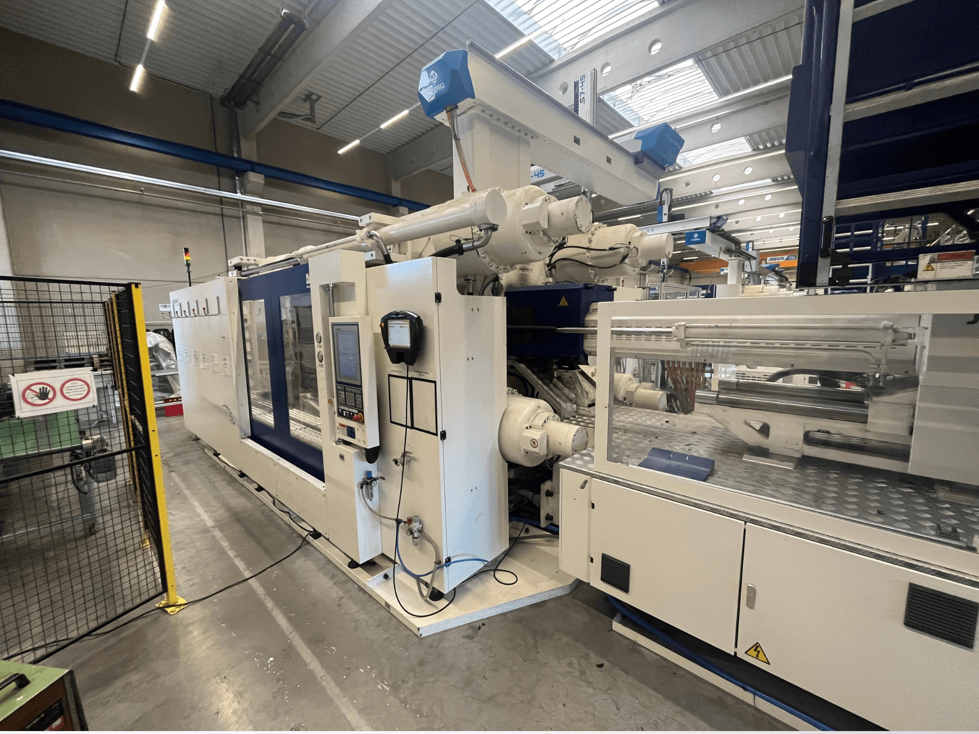 Macchina per lo stampaggio a iniezione in bianco e blu, vista frontale, con pannello di controllo, braccio robotico e segnaletica di sicurezza.