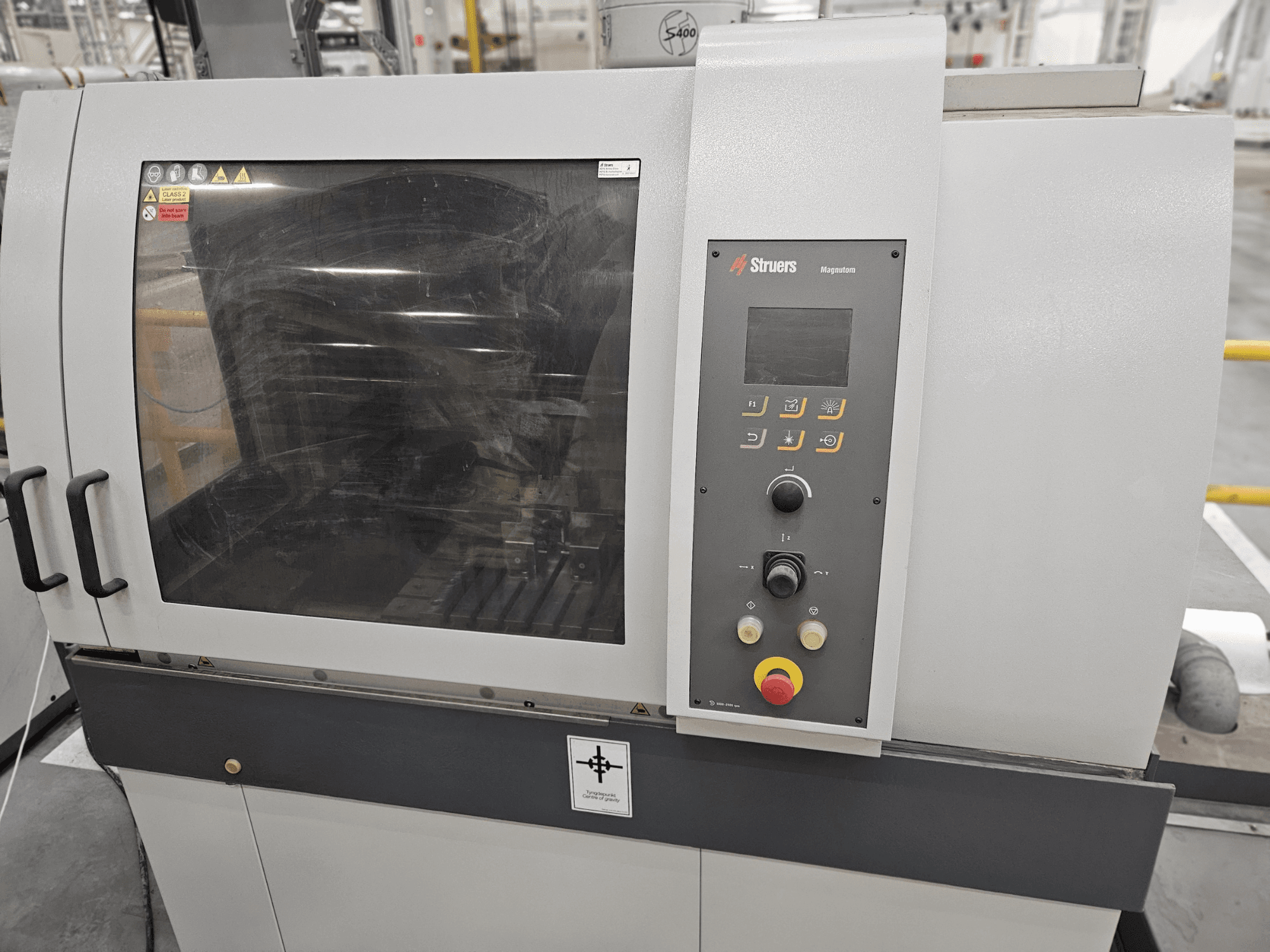 Vista frontale della macchina Struers Magpentor con pannello di controllo, pulsanti e un'ampia finestra di visualizzazione. Etichette tecniche visibili.