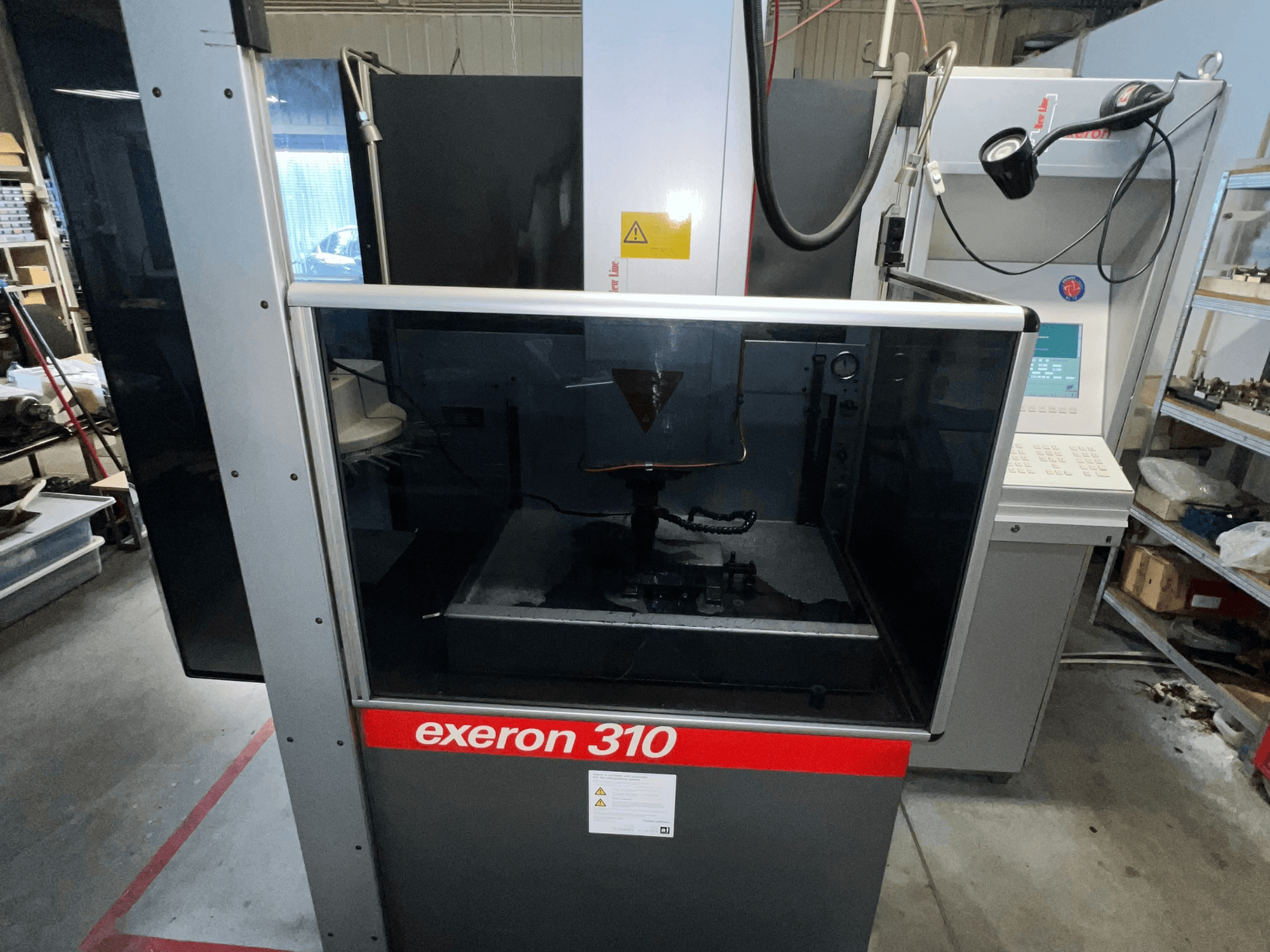 Vista frontale di una macchina Exeron 310, che mostra la camera di vetro e il pannello di controllo accanto alla scaffalatura della postazione di lavoro.