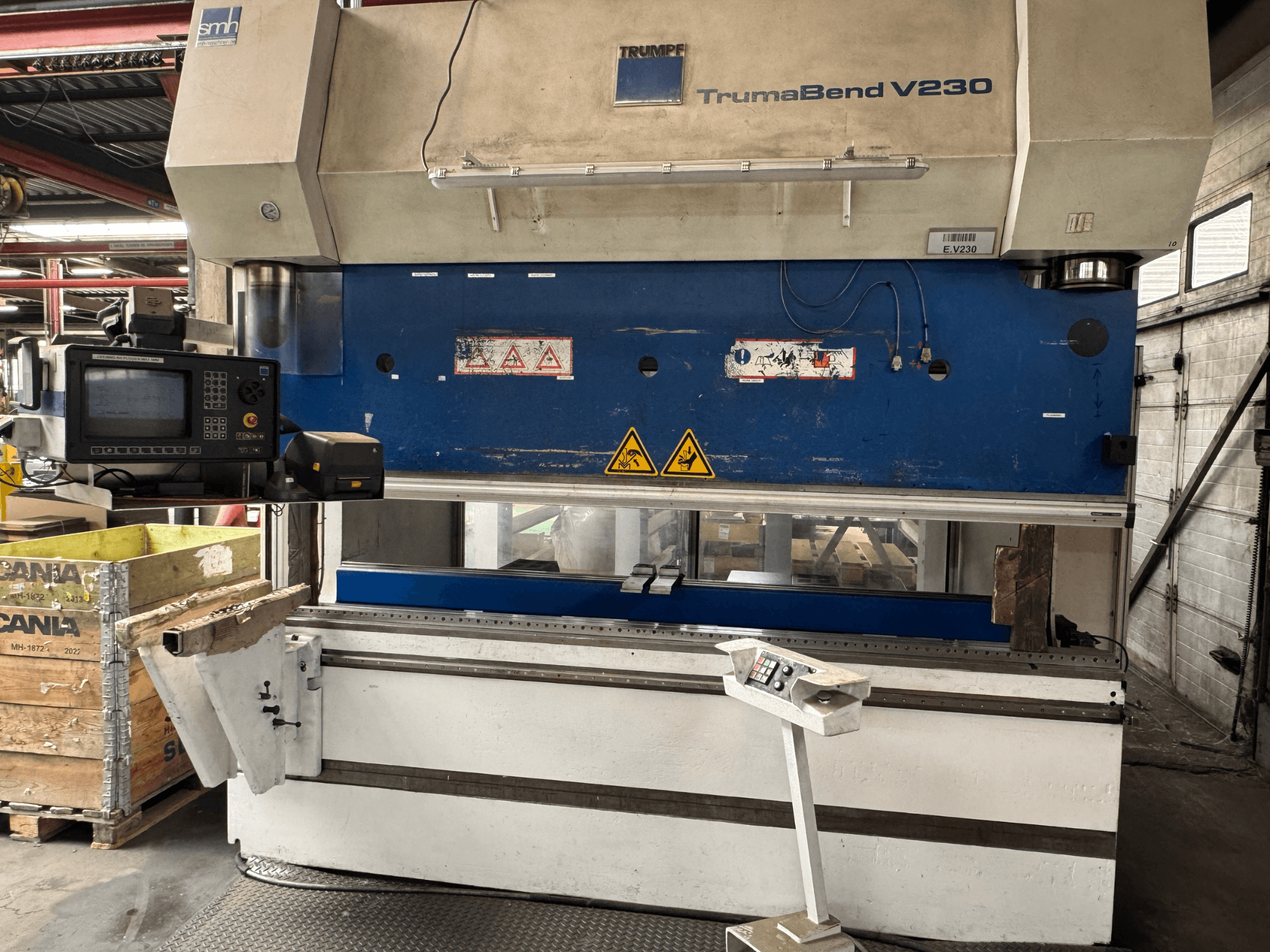 Pressa piegatrice TRUMPF TrumaBend V230, vista frontale con pannello di controllo, etichette di avvertimento e braccio di supporto del pezzo visibili.