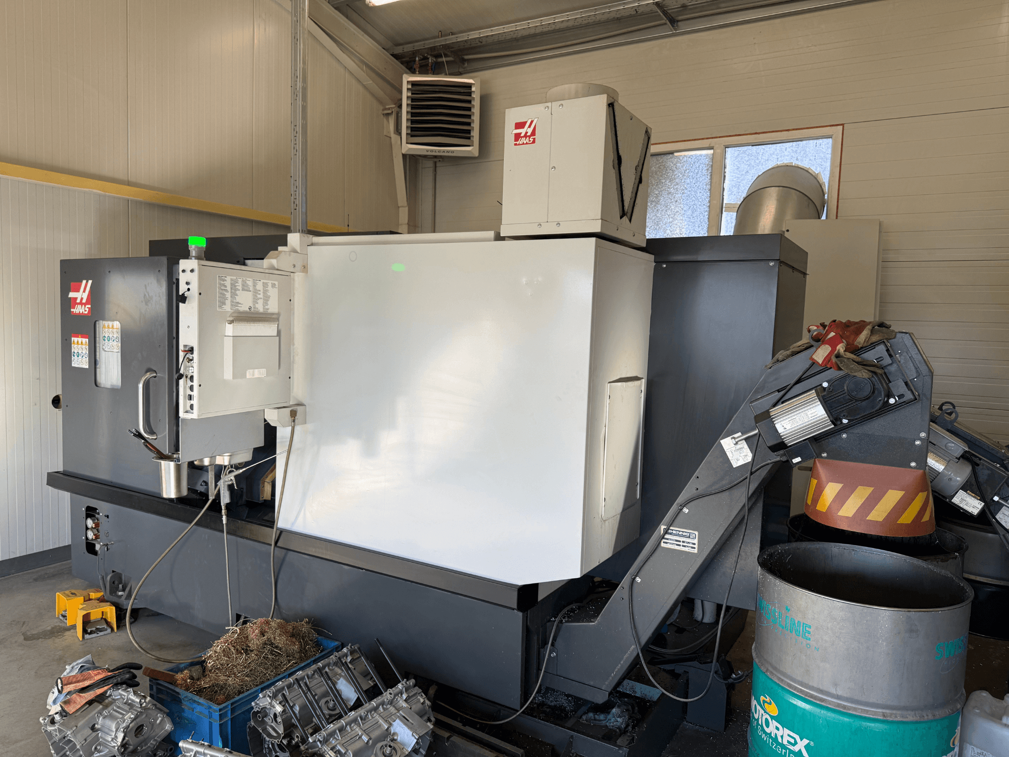 Macchina CNC Haas con pannello di controllo, vista laterale; linee del refrigerante e dell'olio visibili; scarti metallici in un contenitore blu; ambiente industriale.