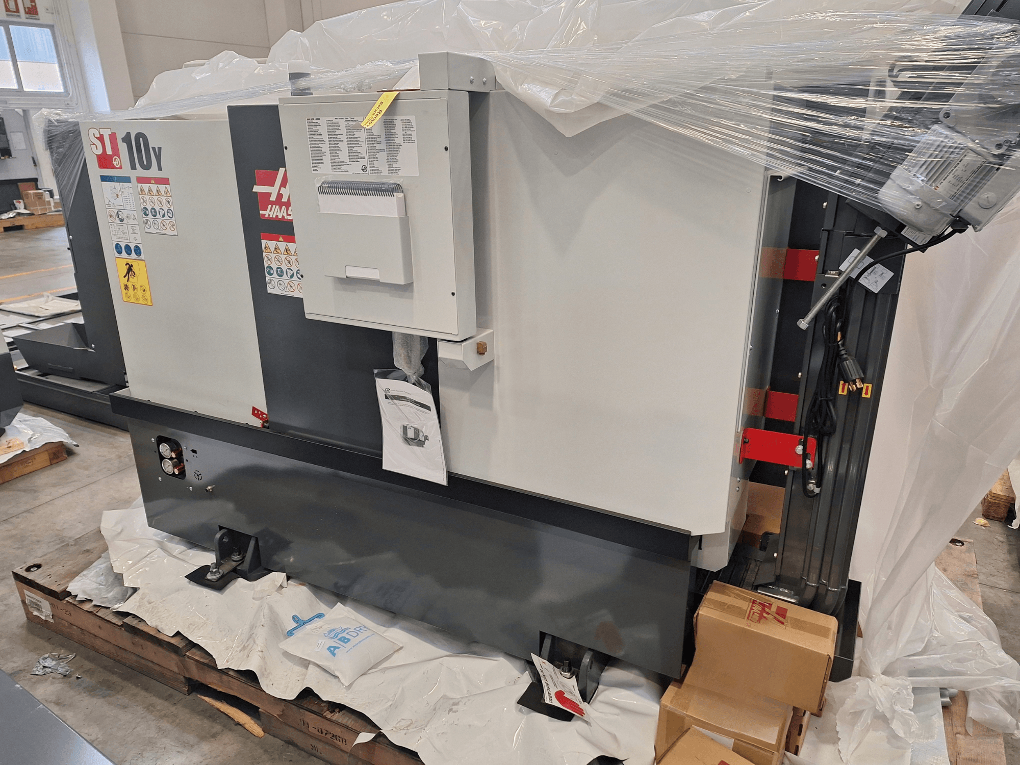 Vista laterale del tornio orizzontale HAAS ST-10Y EU, avvolto nella plastica, con pannello di controllo ed etichette di avvertenza visibili.