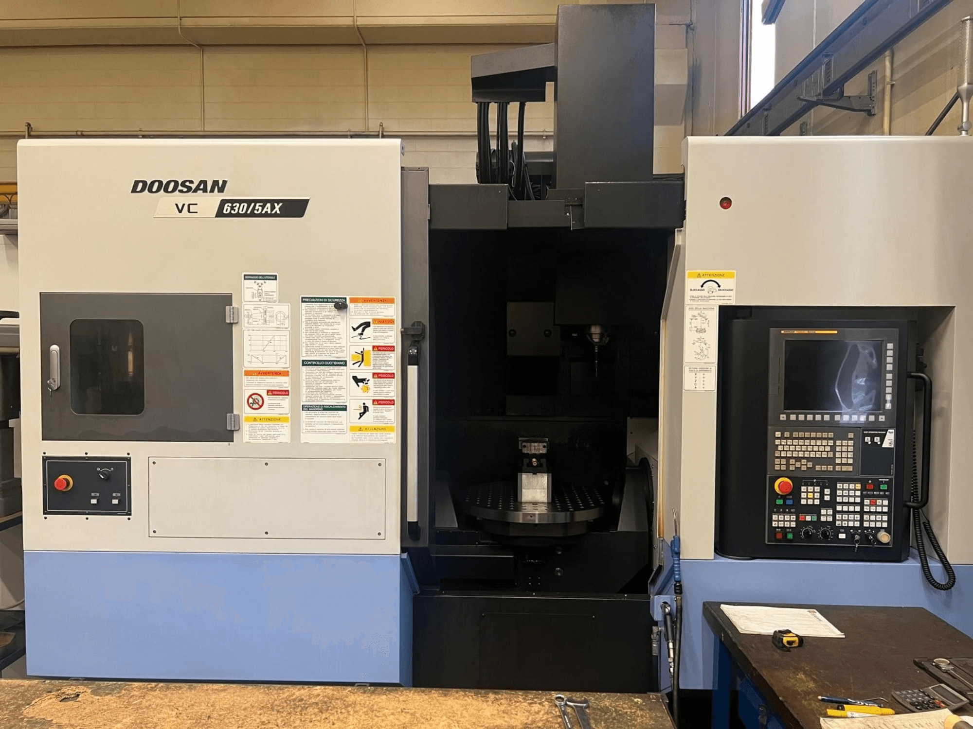 Vista frontale di un centro di lavorazione verticale DOOSAN VC 630/5AX, che mostra il pannello di controllo e l’area del mandrino con le etichette di sicurezza ben visibili.