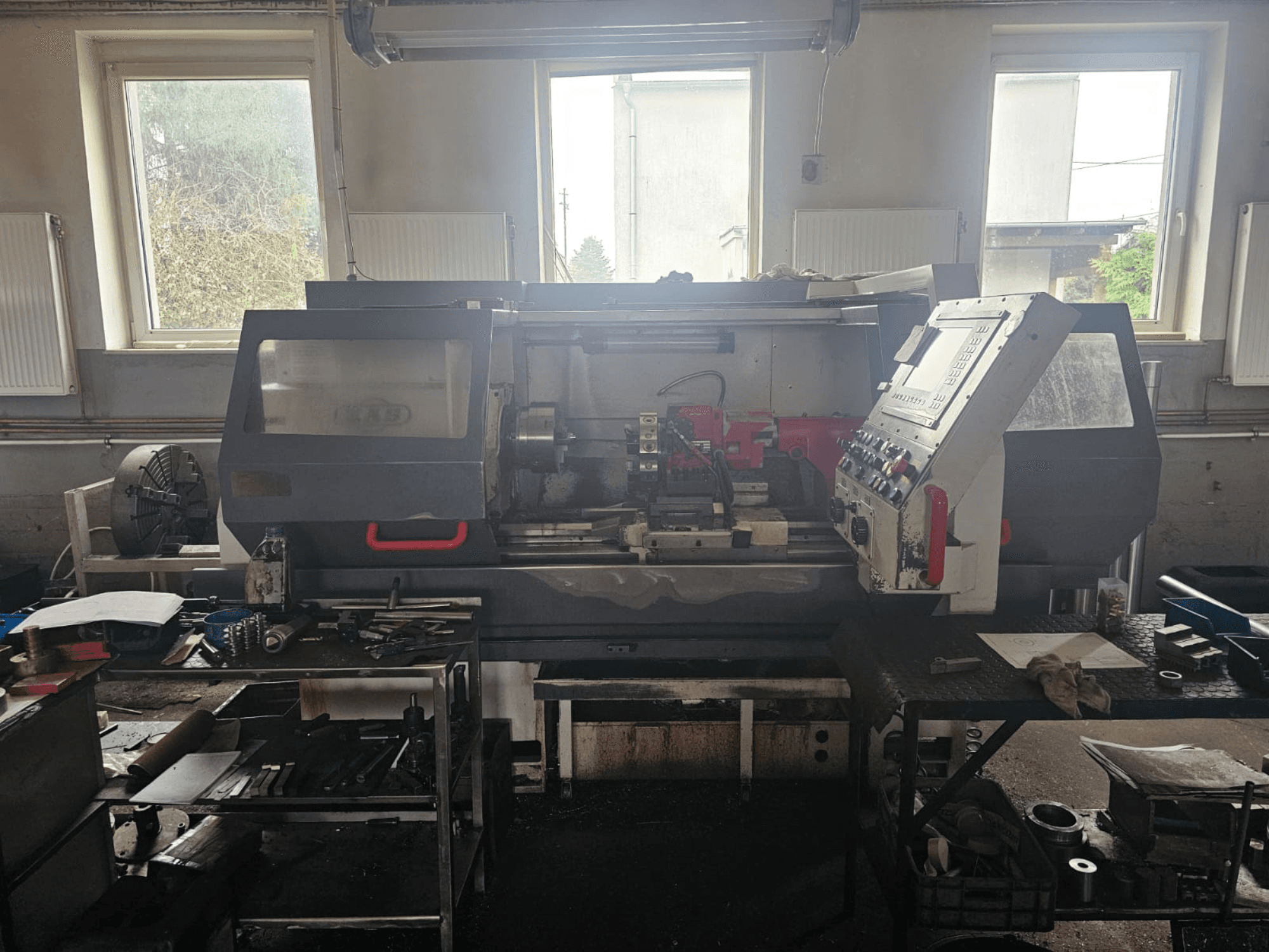 Tornio in un'officina, vista frontale, con pannello di controllo e utensili su tavoli, circondato da finestre e spazio di lavoro disordinato.