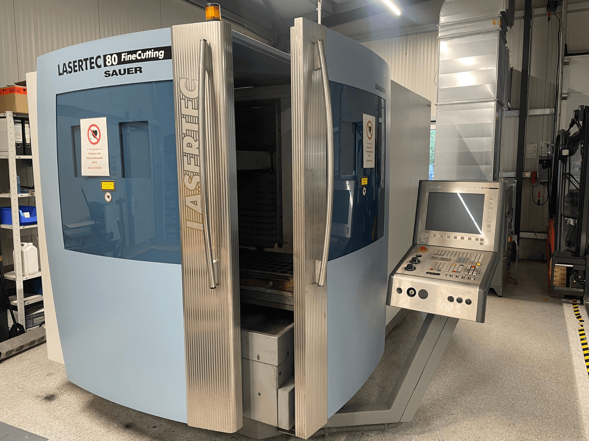 Vista frontale della macchina per il taglio di lamiere DMG Sauer Lasertec80 FineCutting con pannello di controllo, sportelli aperti e segnaletica di sicurezza visibile.