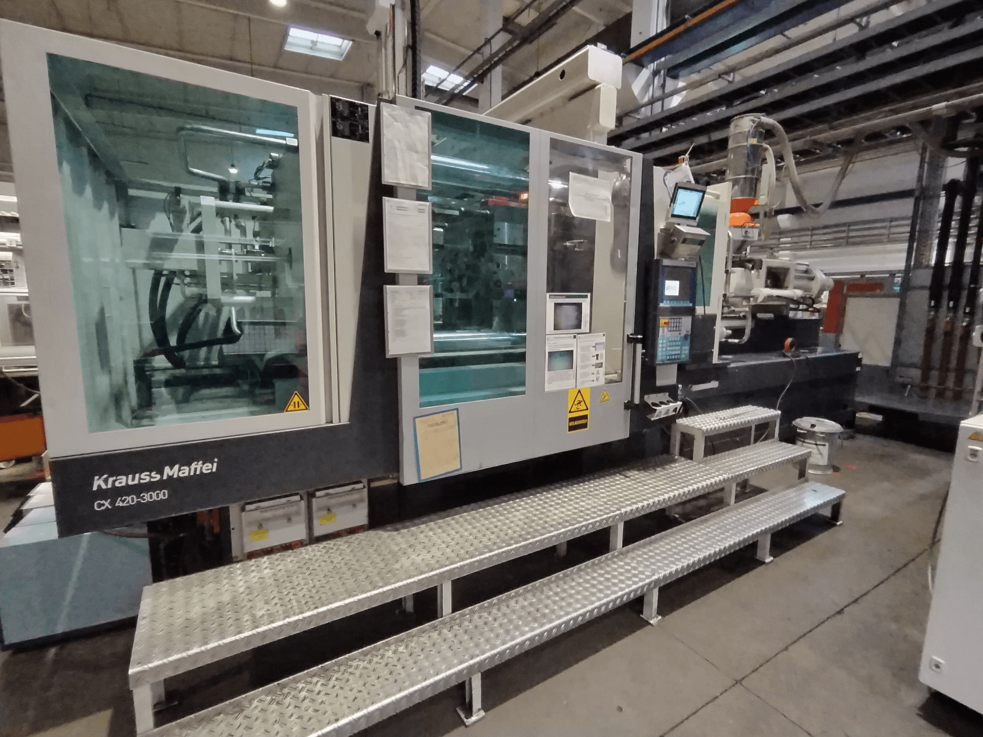 Macchina ad iniezione Krauss Maffei CX 420-3000, vista laterale, con pannello di controllo e segnaletica di sicurezza visibili, posizionata su gradini metallici.