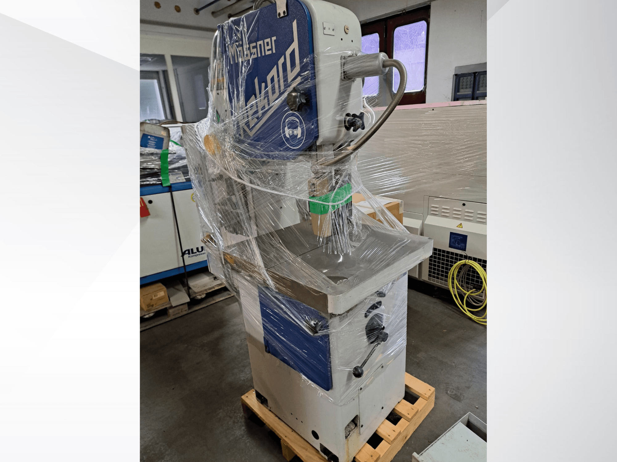 Sega a nastro per metalli MOSSNER Rekord avvolta in plastica; vista laterale che mostra il pannello di controllo e la base su un pallet di legno in un'officina.