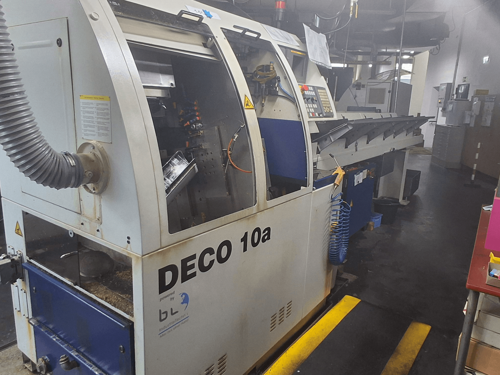 Tornio di tipo svizzero TORNOS DECO 10a, vista frontale; presenta un pannello di controllo, meccanismi visibili e un tubo di aspirazione della polvere.