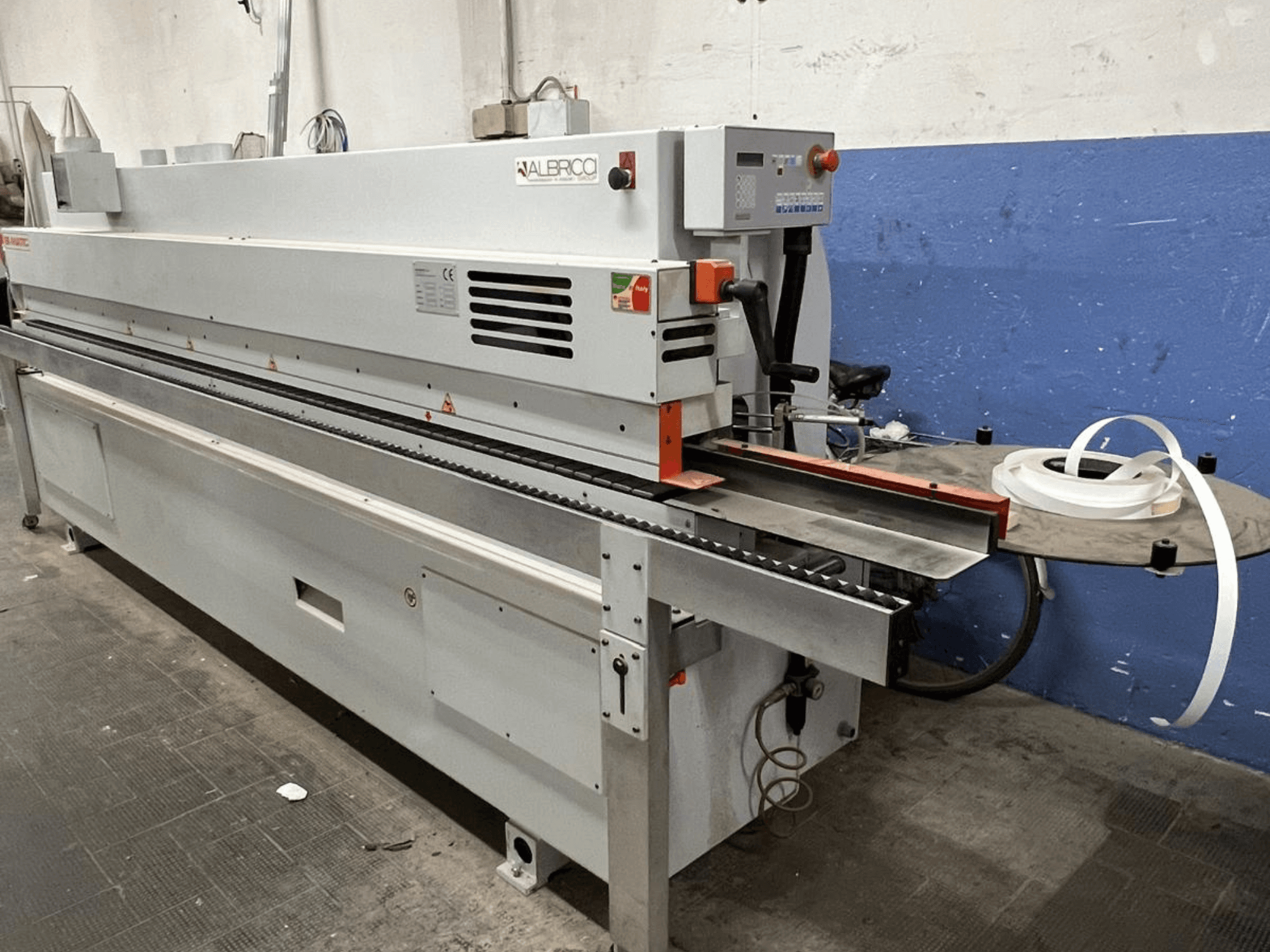 Macchina bordatrice ALBRICO con vista frontale, caratterizzata da un lungo nastro trasportatore e da un rotolo di nastro adesivo collegato.