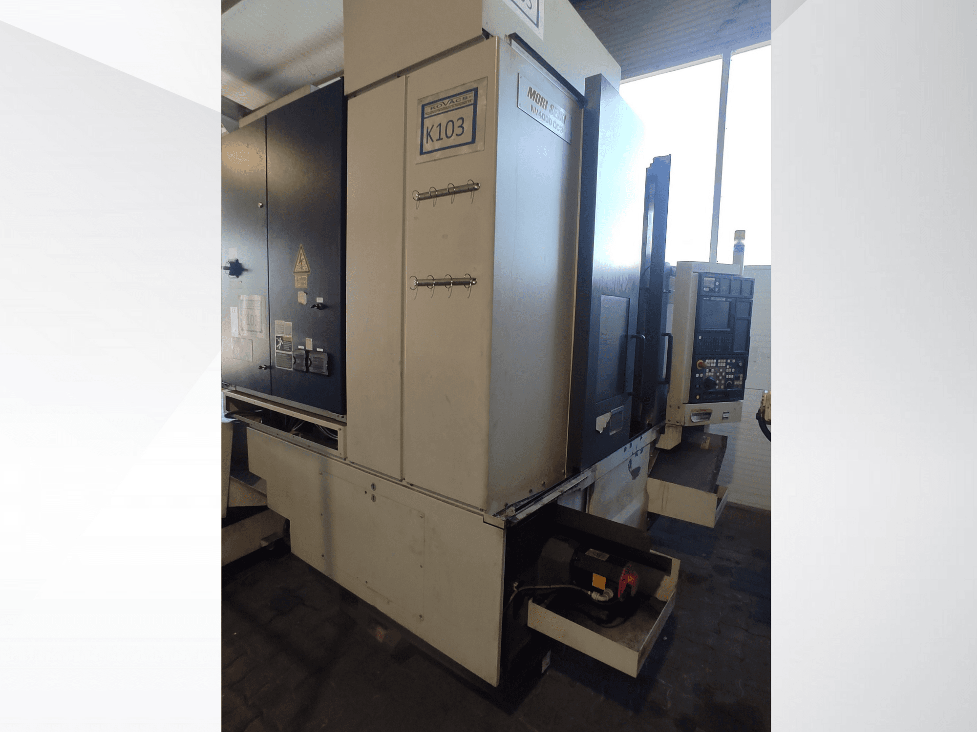 Macchina CNC Mori Seiki K103 in vista frontale, con pannello di controllo, etichette di sicurezza e componenti elettrici visibili.