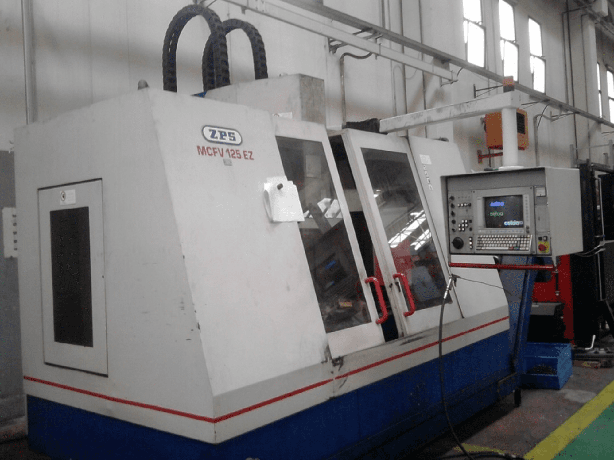 Macchina CNC ZPS MCFV 125 EZ, vista frontale con pannello di controllo e maniglie rosse, in un ambiente di officina con tubi flessibili a vista.