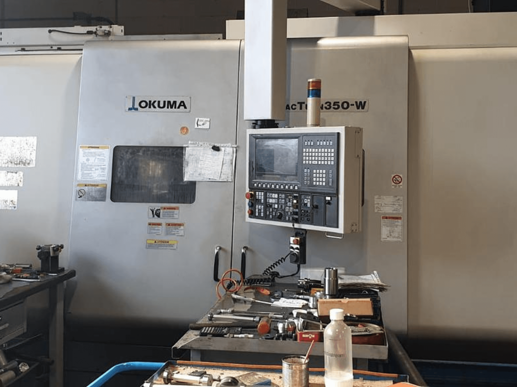 Centro di tornitura Okuma MAC TURN 350-W, vista frontale con pannello di controllo e spazio di lavoro ingombro di utensili e una bottiglia.