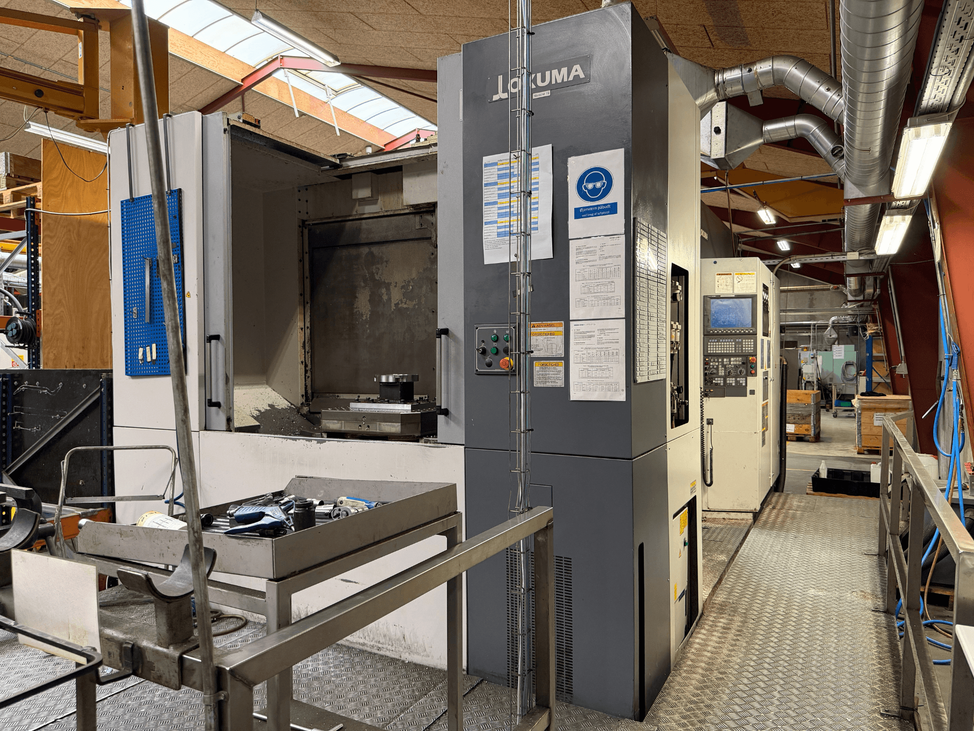 Centro di lavorazione orizzontale Okuma MA-600HB, vista frontale, con gli utensili disposti su un piano di lavoro in metallo e i comandi della macchina.
