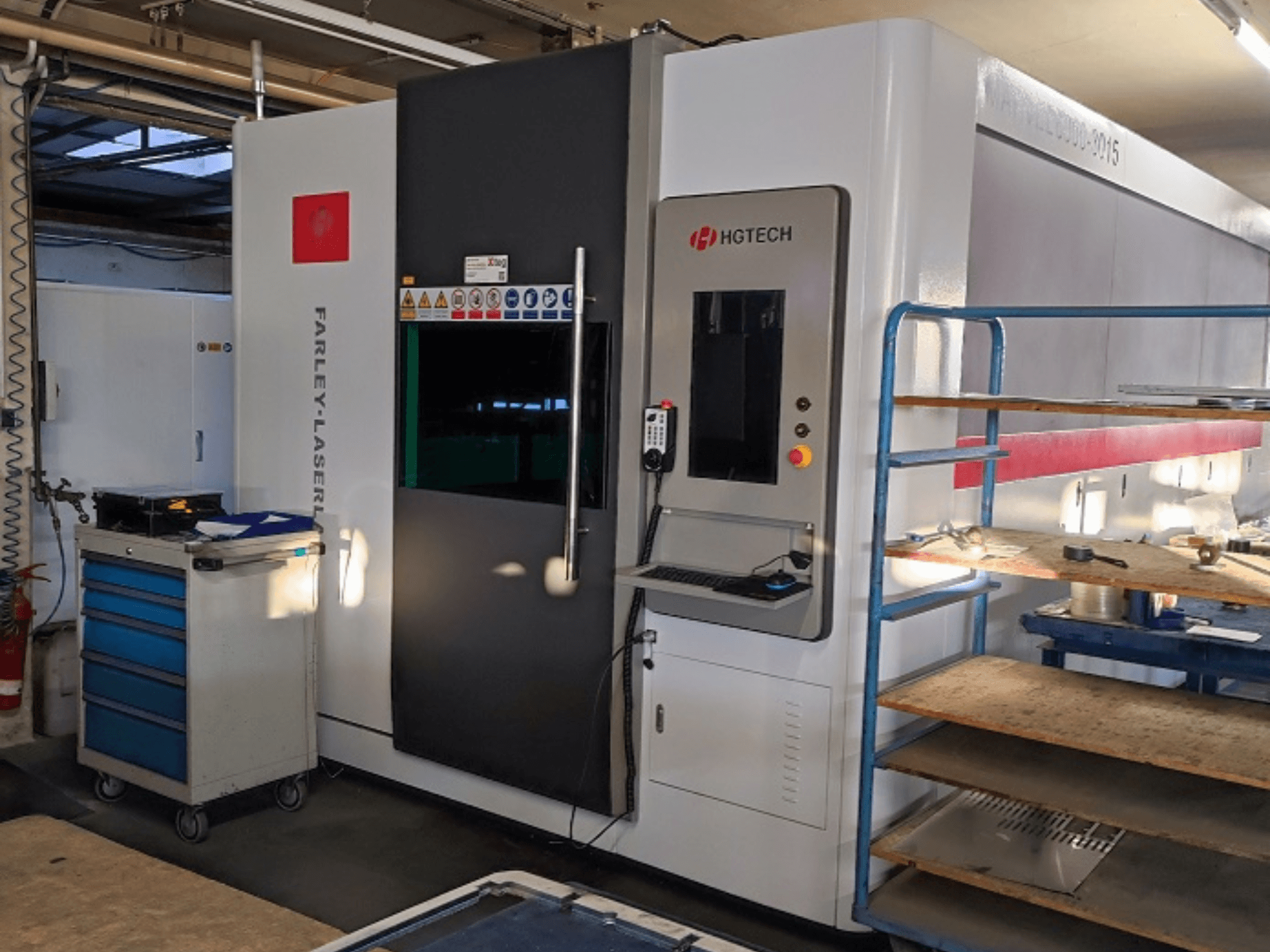 Macchina di taglio laser in fibra HGTECH Marvel 6000-3015, vista frontale con pannello di controllo e magazzino adiacente.