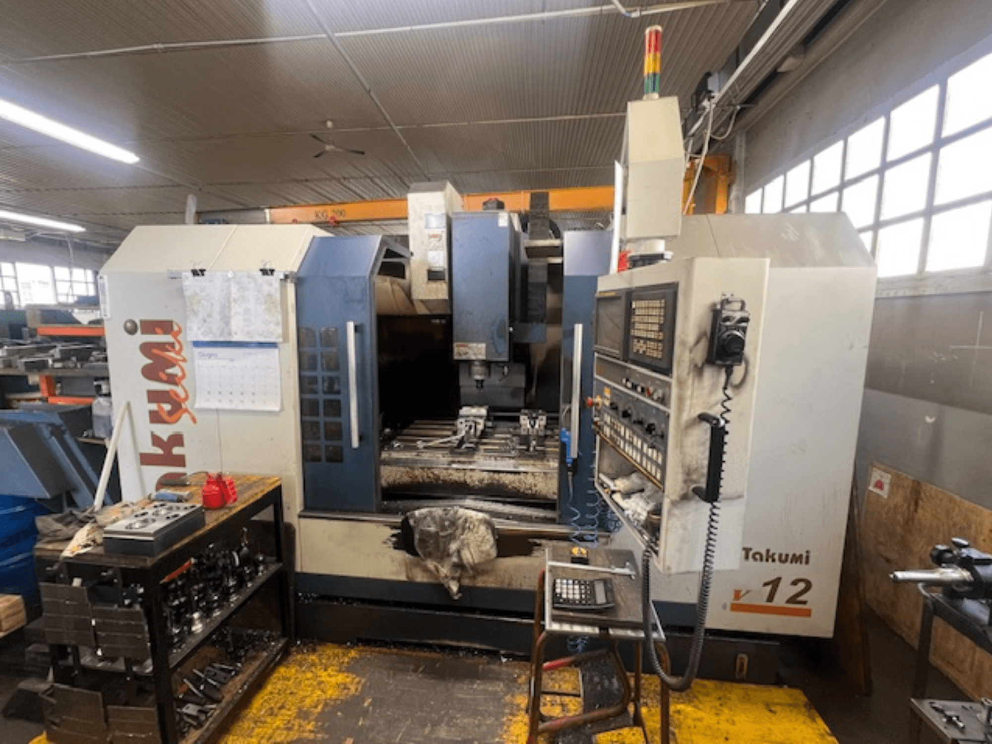 Fresatrice CNC Takumi V12 in vista laterale, con pannello di controllo, area di taglio e utensili sulla postazione di lavoro.
