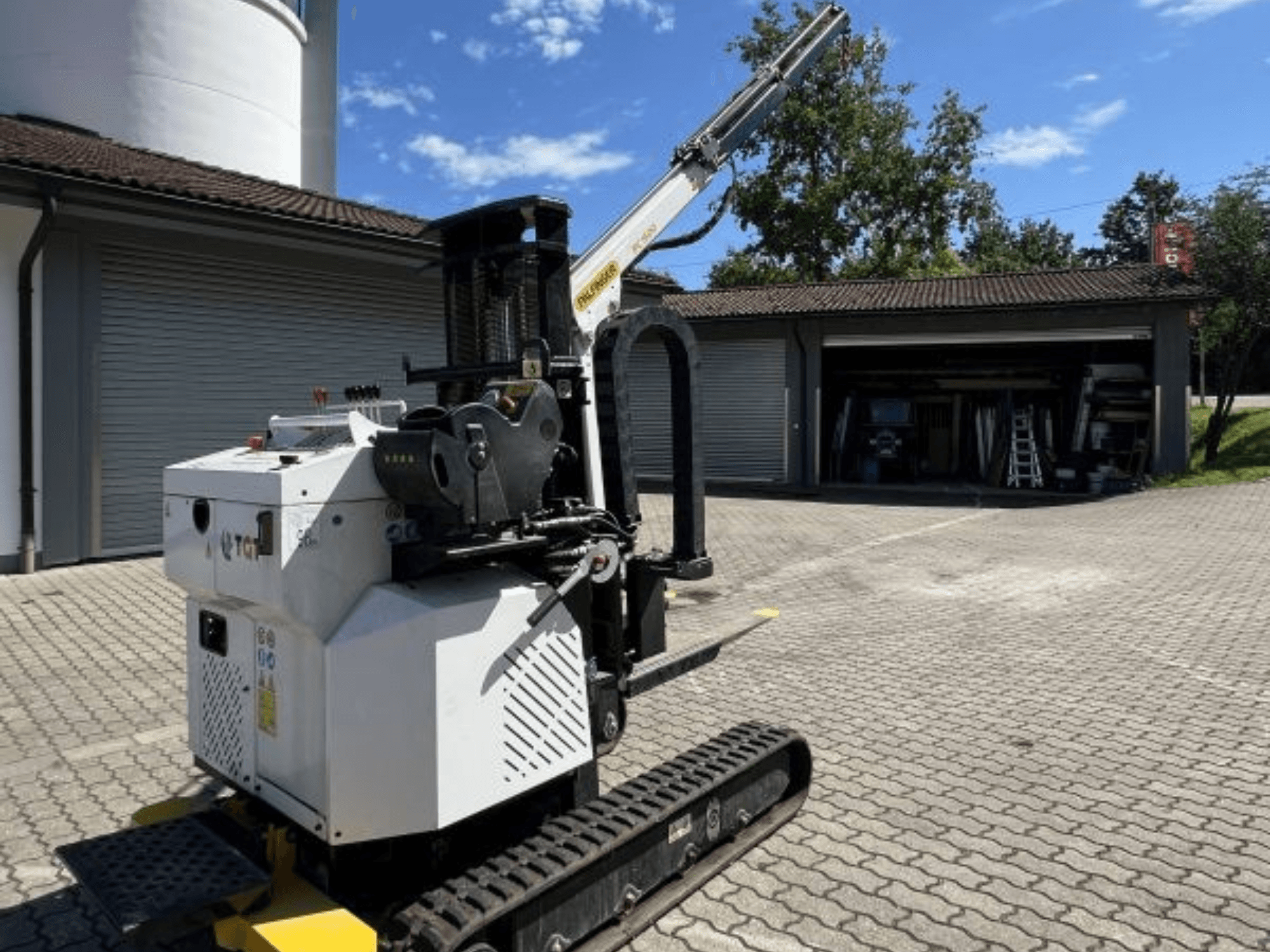 Mini gru da cantiere TGI, vista laterale, che mostra i cingoli e il braccio montato, parcheggiata su una superficie di mattoni presso un garage.