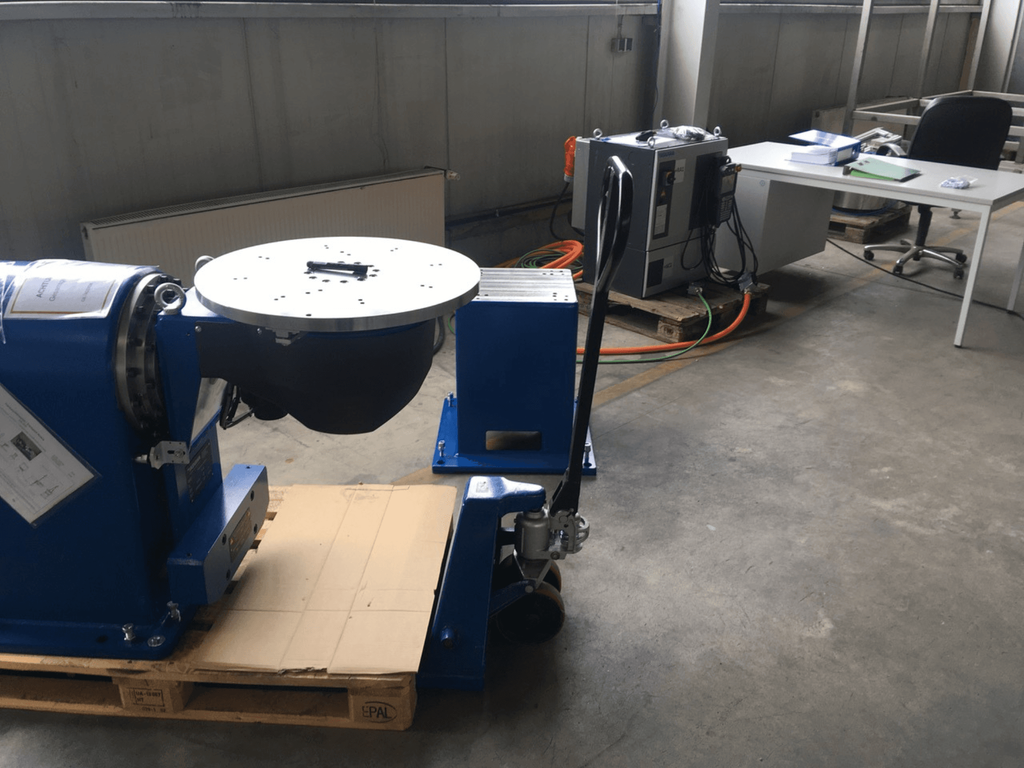 Apparecchiatura industriale blu con base circolare e pannello di controllo, vista dal davanti, su un pallet di legno in un ufficio.