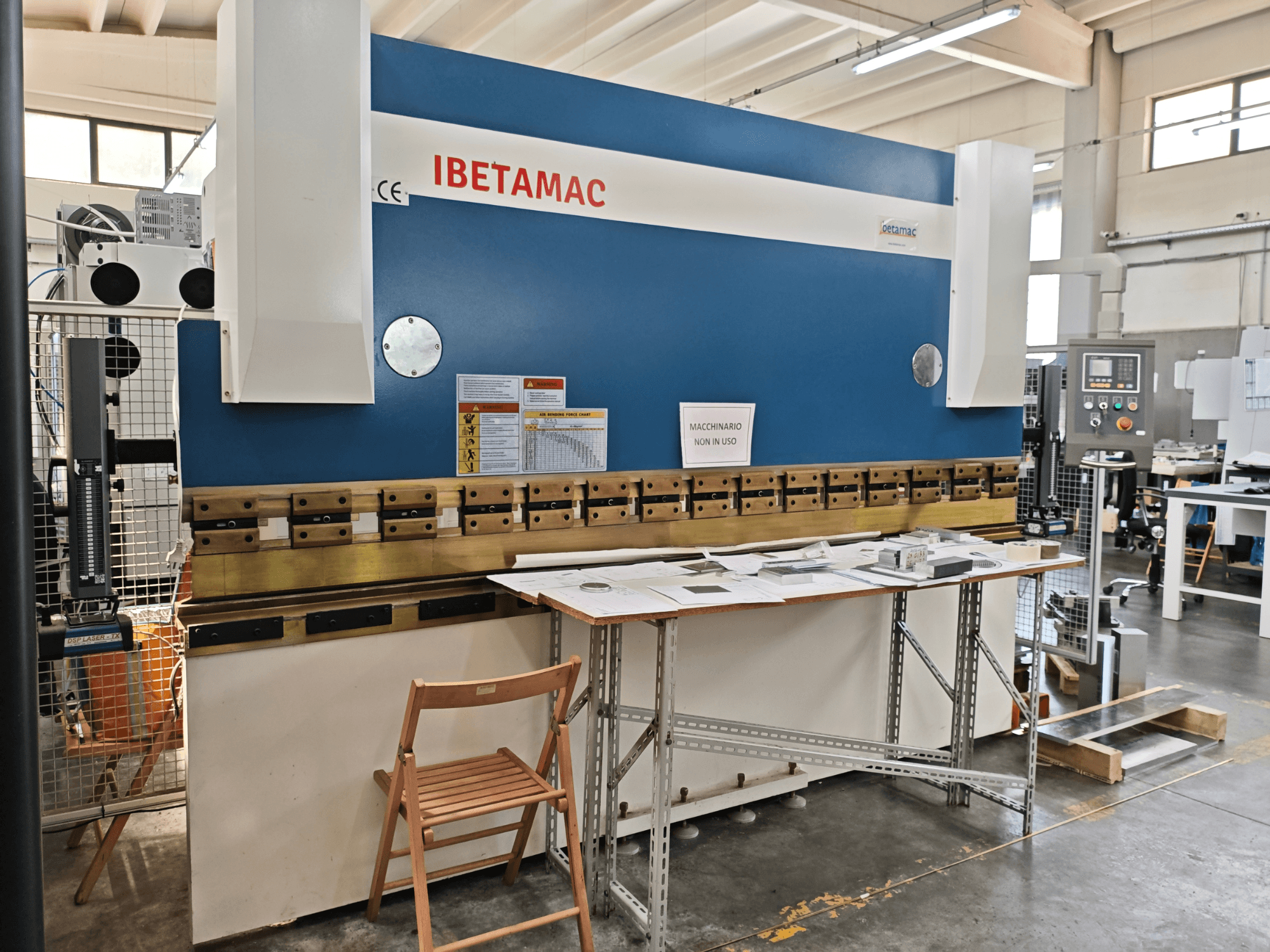Piegatrice IBETAMAC 3200 100T in un'officina; vista laterale che mostra il pannello di controllo, l'area di lavoro e le avvertenze di sicurezza.