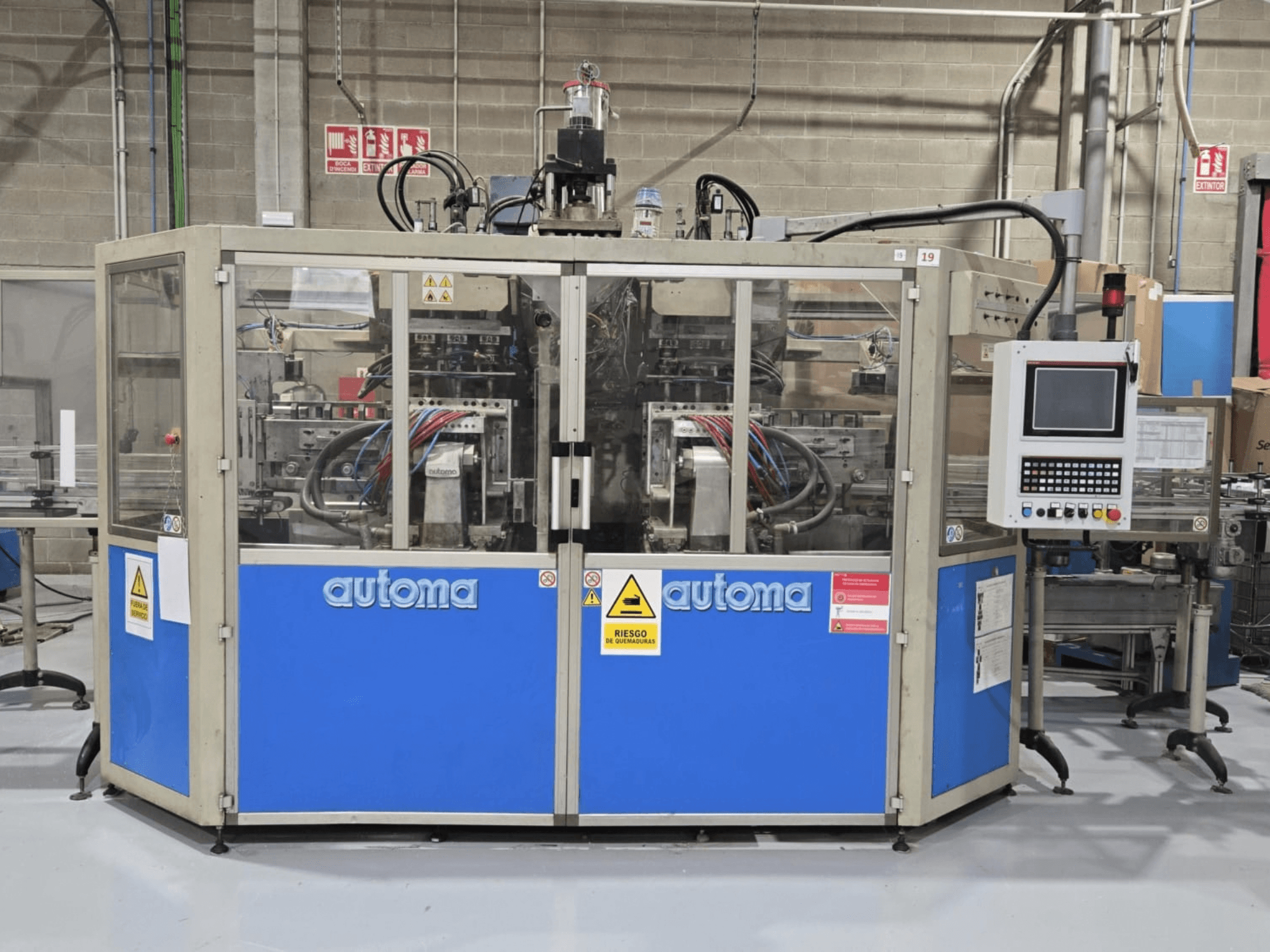 Vista frontale della macchina per lo stampaggio a soffiaggio AUTOMA APEX AT500D, con pannello di controllo e vari tubi flessibili, collocata in un ambiente industriale.