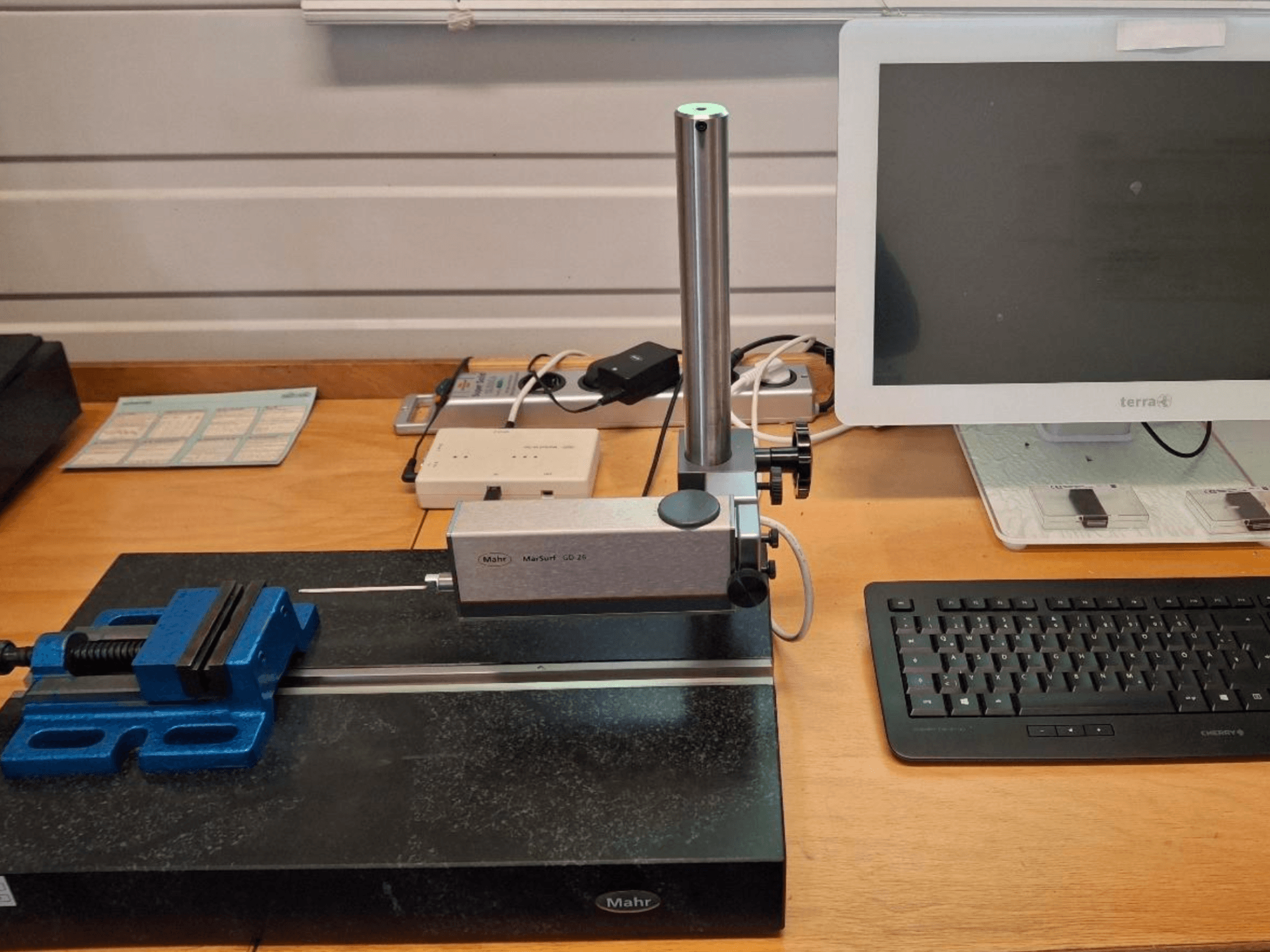 Macchina di misura MAHR MARSURF GD 26 su un banco di lavoro, con sonda di misura, morsa blu e monitor del computer. Vista frontale.
